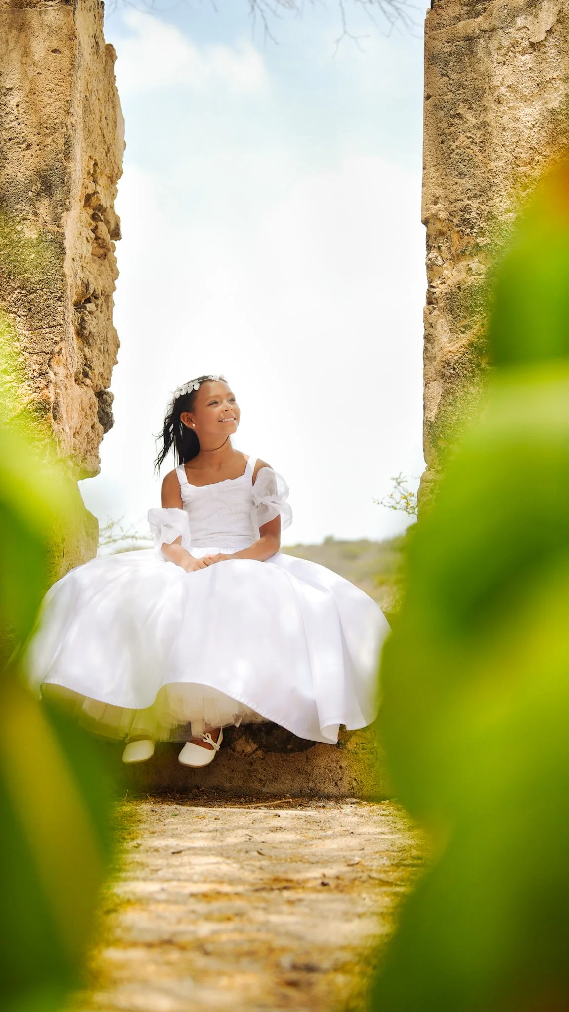 Young girl sitting outdoors, dressed in a white dress with puffy sleeves and a flower crown, framedaruba goldmine landscape, smiling and looking off into the distance.