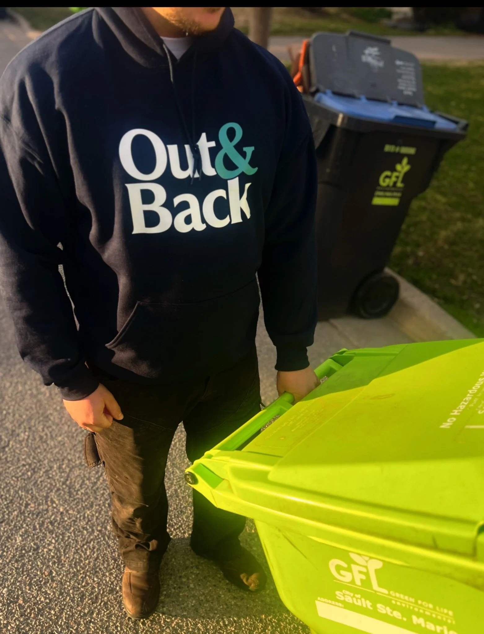 Person in black hoodie with the words 'Out & Back' in white and teal, standing near green recycling bin labeled 'GF Recycling' at curbside.
