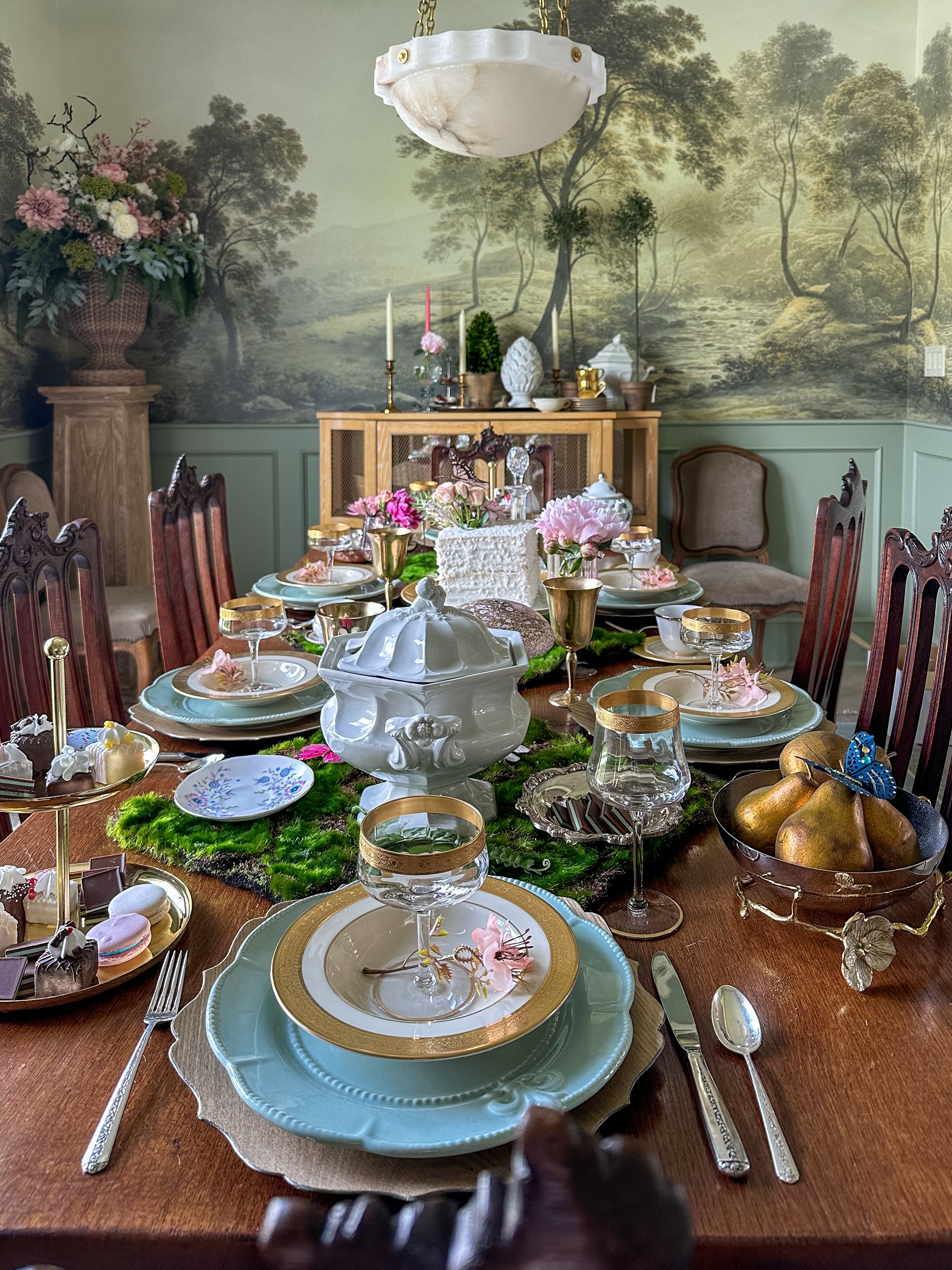Editorial dining tablescape styled by Melissa B. Rodgers featuring layered vintage china, gold accents, florals,moss runner, and dramatic mural wallpaper.