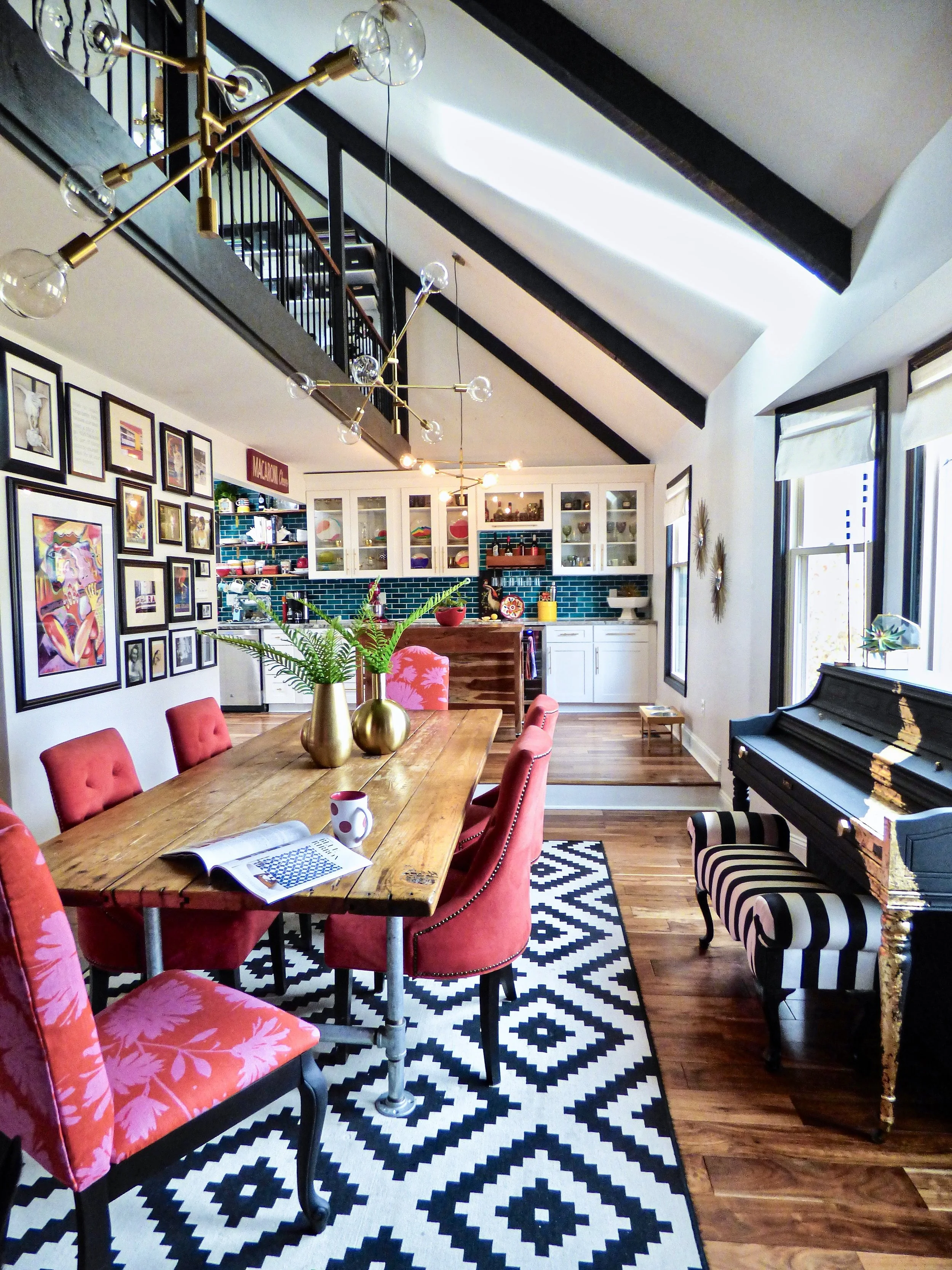 Eclectic maximalist dining room with black ceiling beams, pink and red upholstered dining chairs, graphic black and white rug, gallery wall and open kitchen shelving and teal kitchen backsplash designed by Melissa B. Rodgers