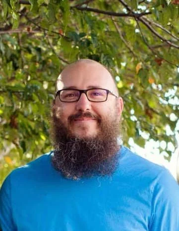 Man with glasses and a beard smiling outdoors with trees and green leaves in the background.