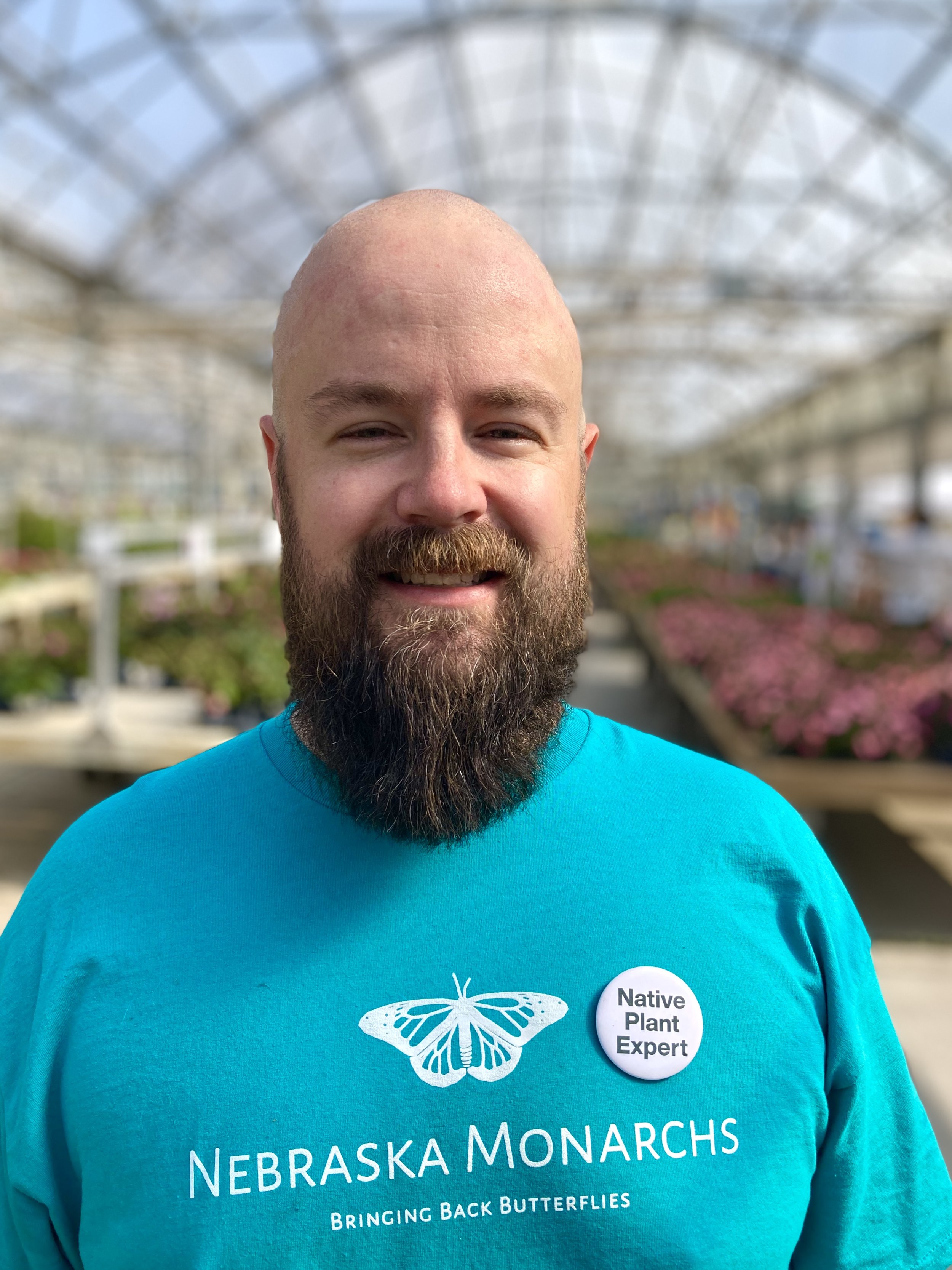 A man with a beard and bald head smiling in a greenhouse. He is wearing a blue t-shirt with a monarch butterfly graphic and a button that says "Native Plant Expert". The background has blurred plants and flowers.