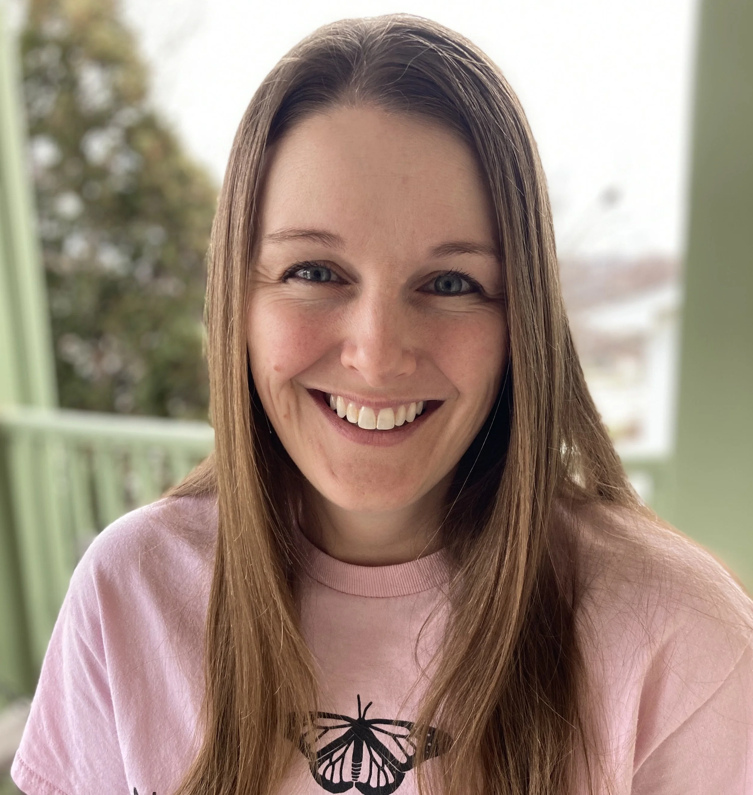 Close-up of woman smiling with long light brown hair wearing a pink shirt with a butterfly design, standing outdoors on a bright day.