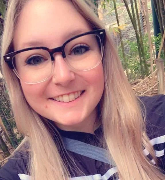 A smiling young woman with long blonde hair and black glasses, outdoors in a lush, green area with bamboo plants in the background.