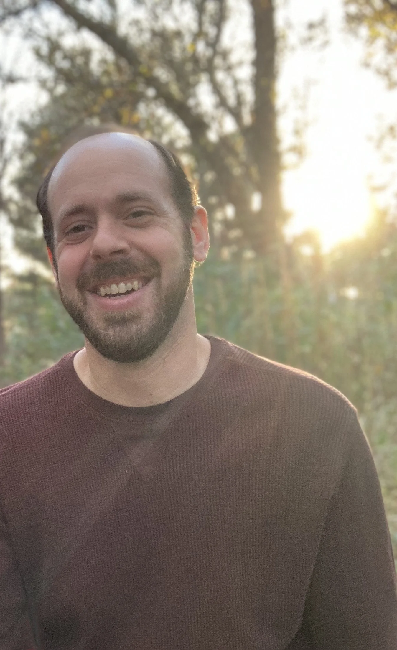 A man with a beard smiling outdoors during sunset, wearing a brown shirt, with trees and sunlight in the background.
