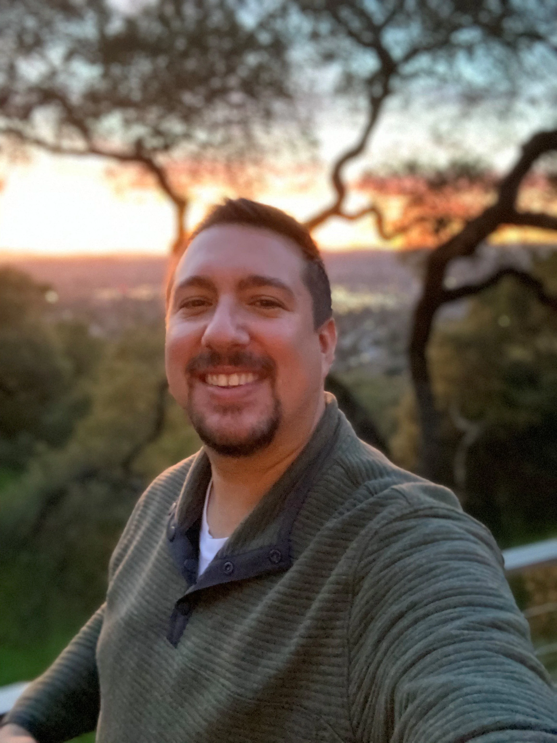 A man with short dark hair and a beard, smiling, taking a selfie in front of a blurred background of trees and sunset.