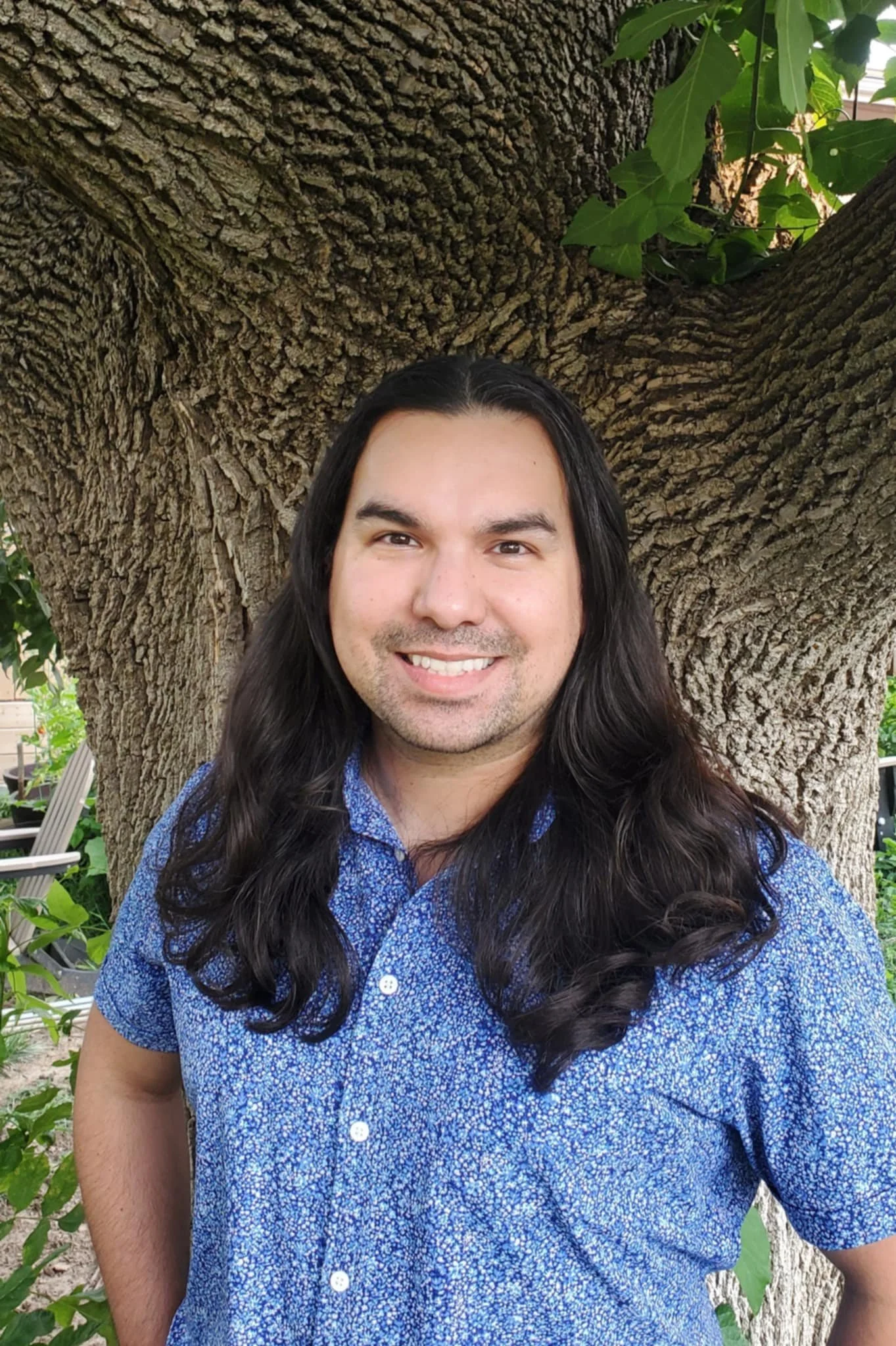 A man with long, dark hair and a light beard, smiling, standing outdoors in front of a large tree with green leaves.