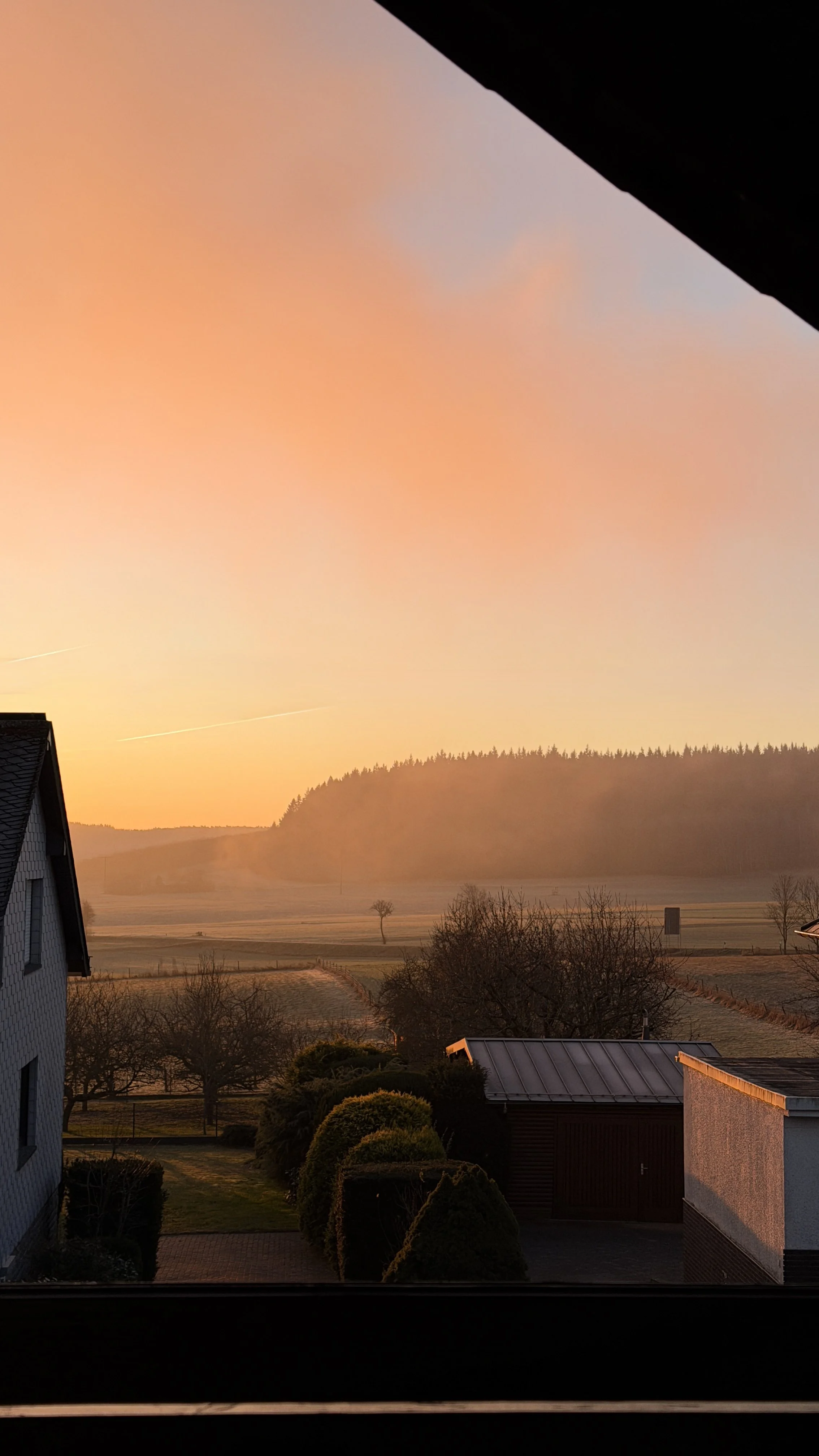Blick auf einen Sonnenaufgang über einem ländlichen Gebiet mit Bäumen, Feldern und Hügeln im Hintergrund, durch ein Fenster gesehen.