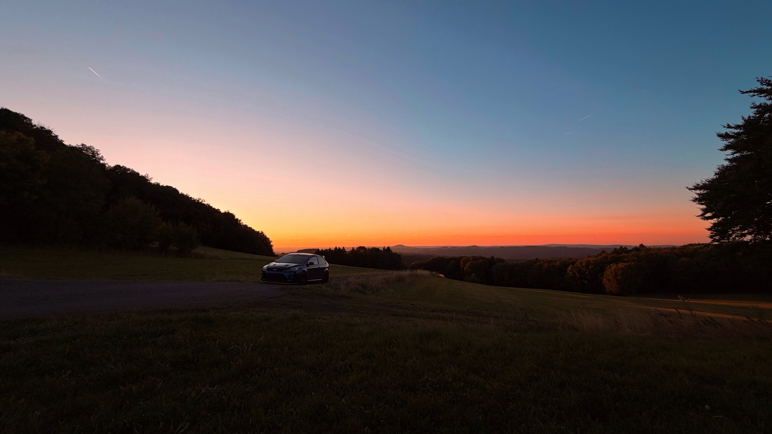 Auto auf einer Landstraße bei Sonnenuntergang, umgeben von Bäumen und hügeliger Landschaft.
