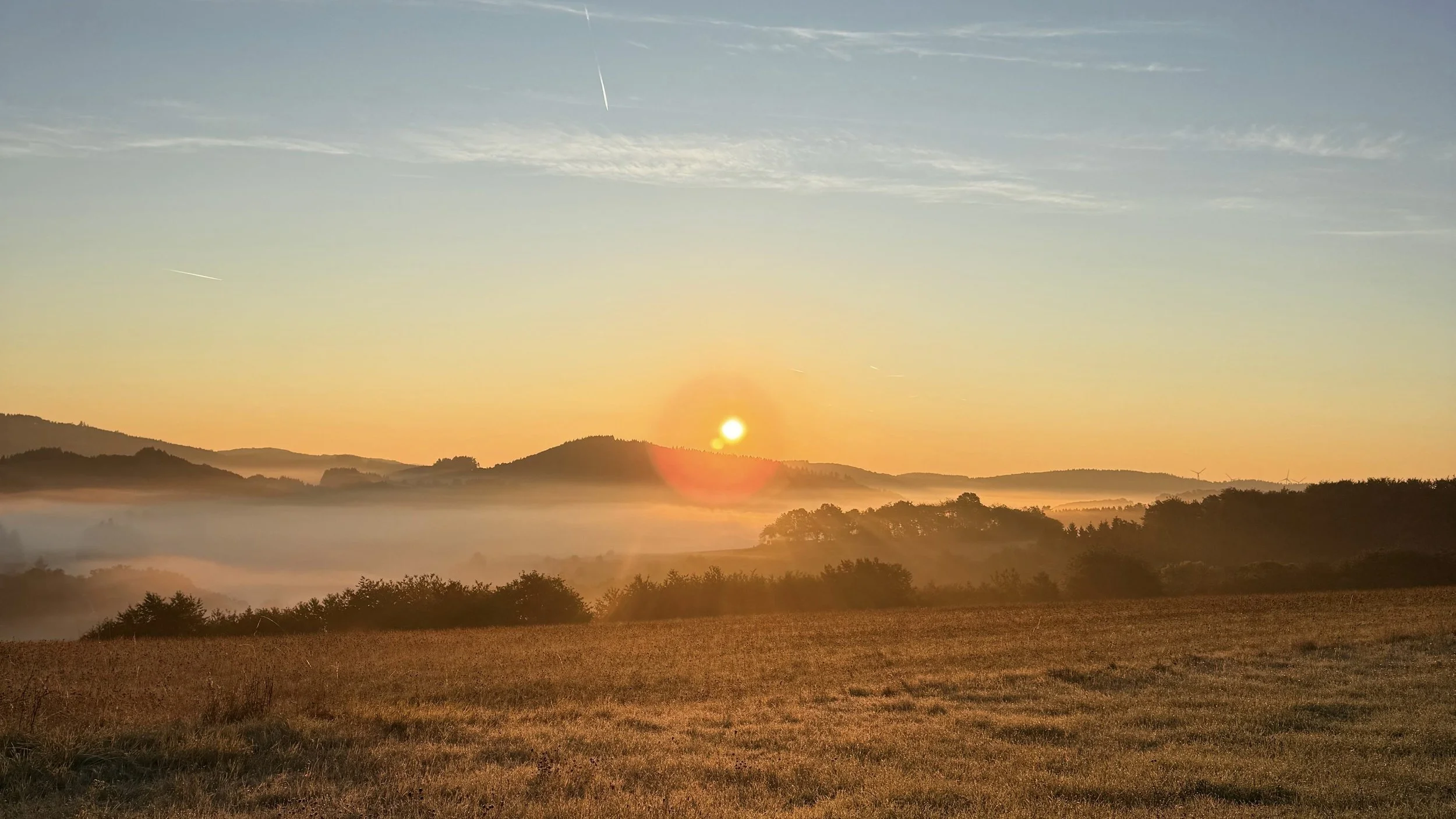 Sonnenaufgang über einem hügeligen Landschaftsbild mit Nebel im Tal und weiten Feldern im Vordergrund.