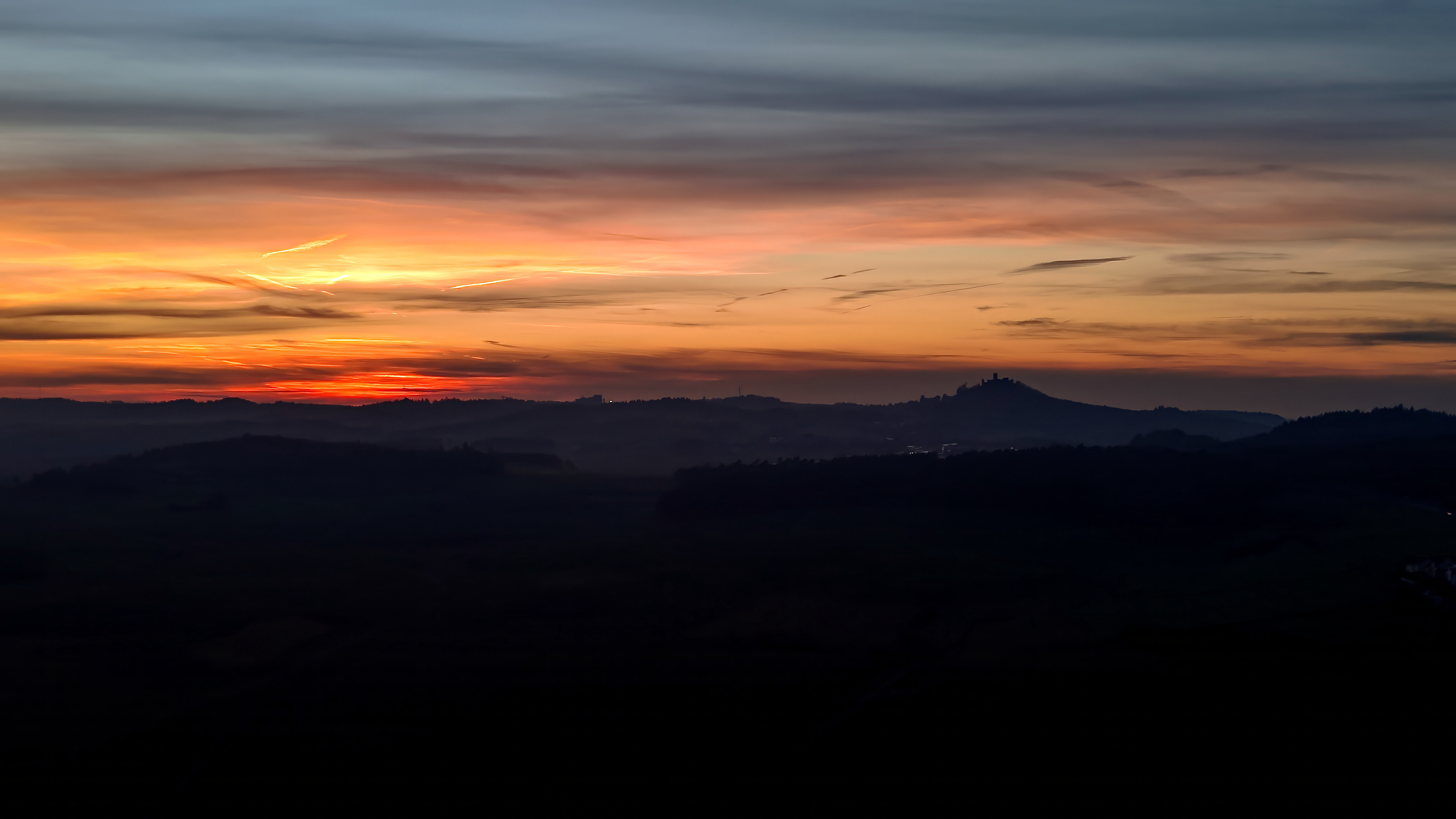 Blick auf eine Berglandschaft bei Sonnenuntergang mit buntem Himmel und Silhouetten von Hügeln im Vordergrund.