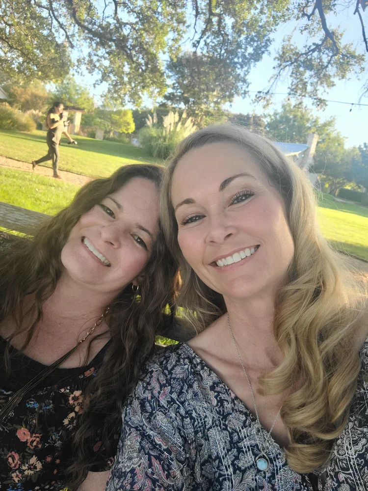Two women smiling outdoors in a park during the daytime with trees and grass in the background. One woman has long wavy hair and the other has slightly curled hair.