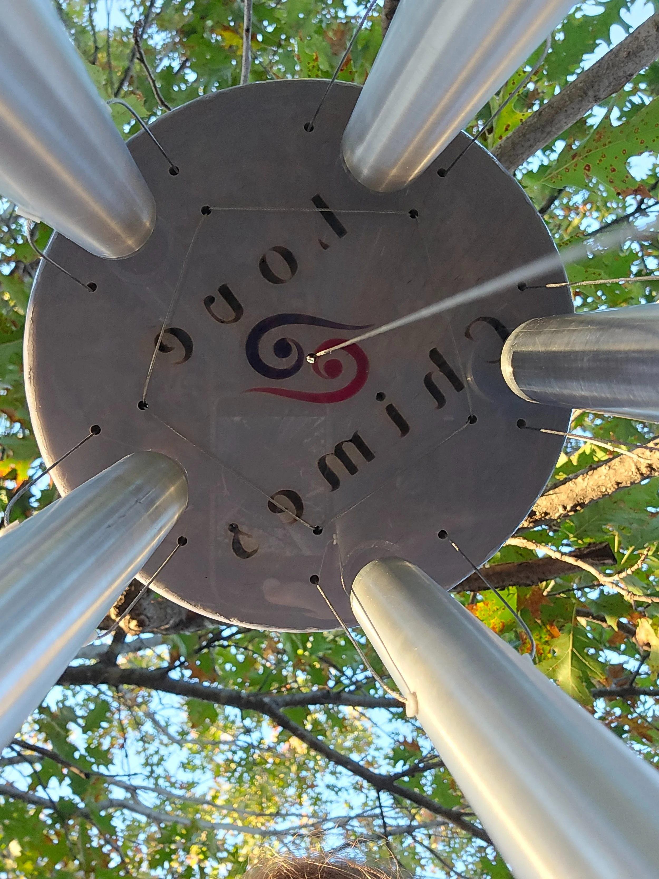 Looking up at a wind chime hanging from a tree with green leaves, showing the bottom of the wind chime, which is circular with cut-out letters and metal tubes hanging from it.