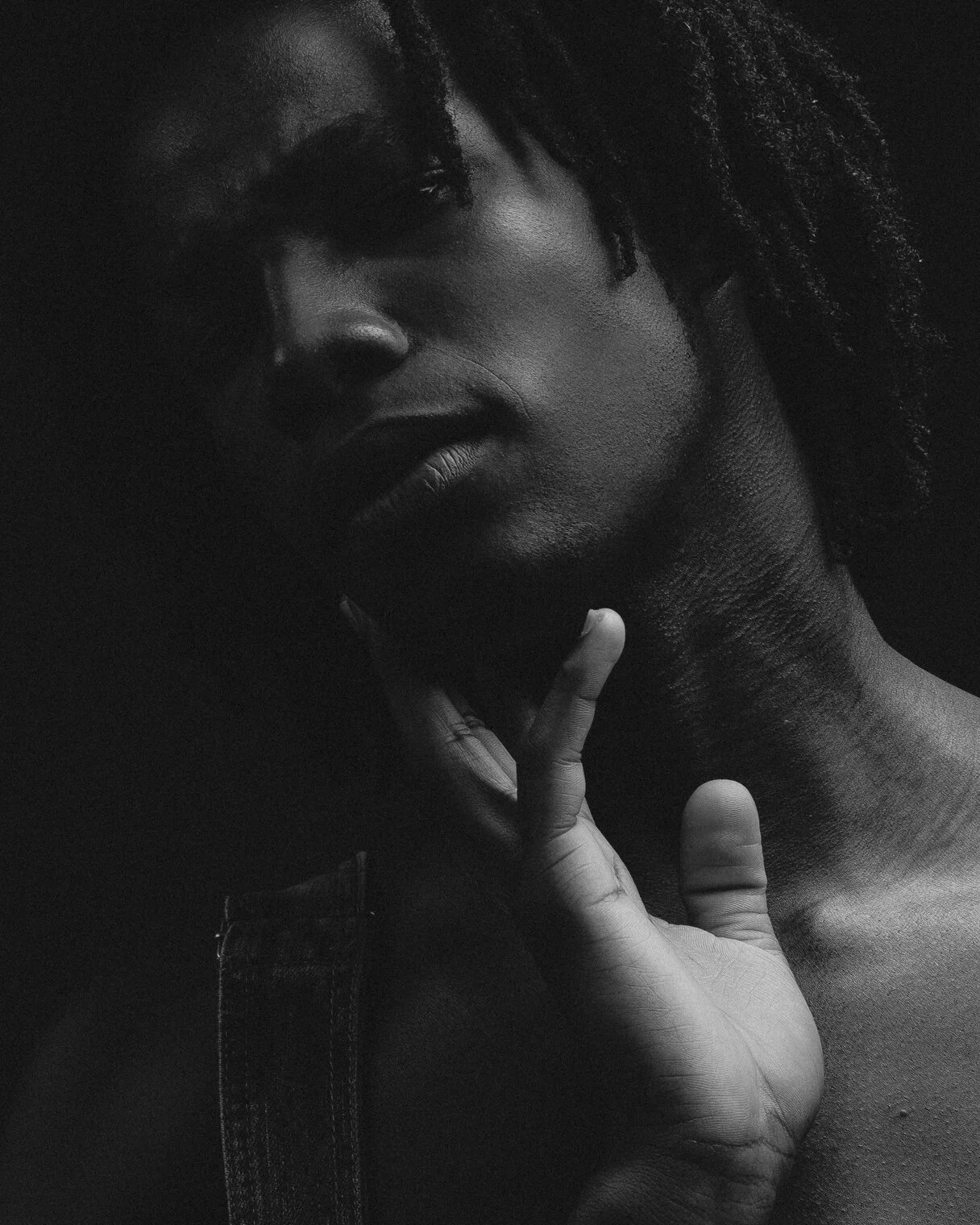 Close-up black and white portrait of a person with dreadlocks, holding their chin with one hand, looking at the camera, with a serious expression.