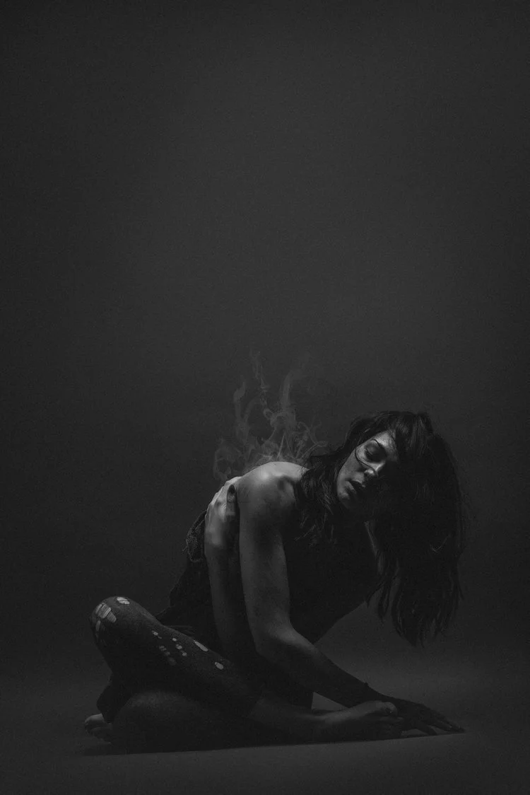 Black-and-white photo of a woman with long hair sitting on the floor, looking down with a contemplative expression, surrounded by a dark background.