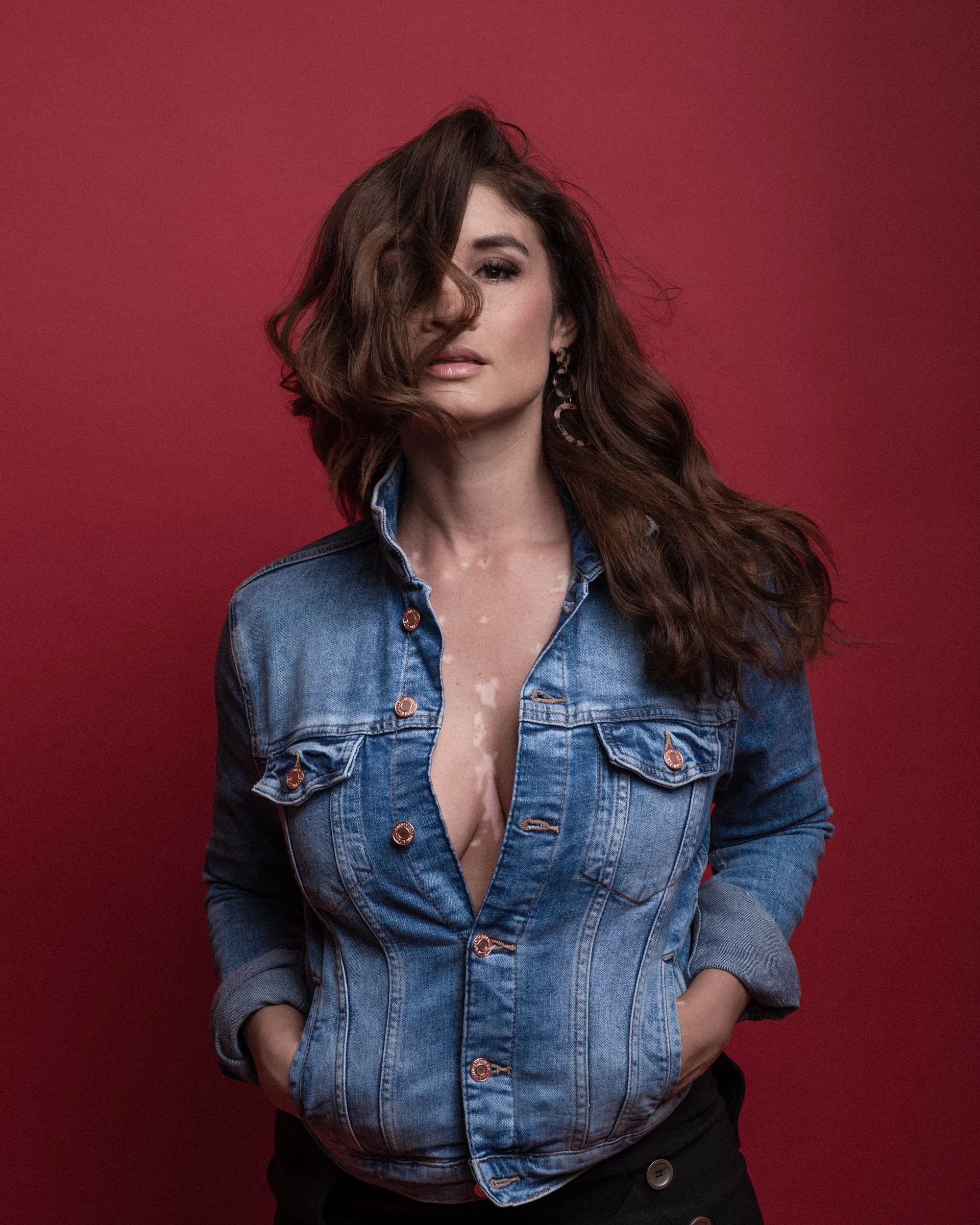 Woman with brown hair, wearing a denim jacket, standing in front of a red background.