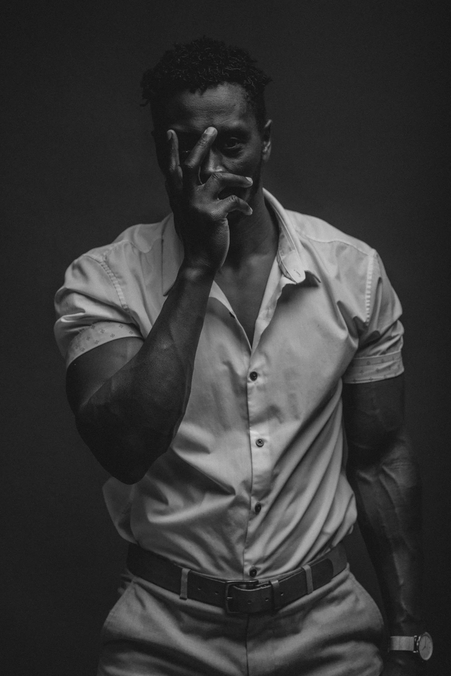 Black and white portrait of a man wearing a light-colored shirt with rolled-up sleeves, adjusting his glasses with his right hand, against a dark background.