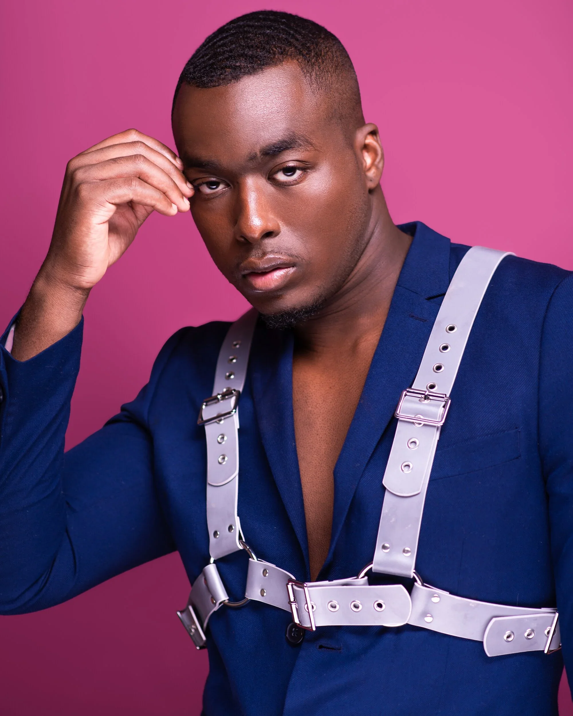 A man with short hair and a serious expression wearing a dark blue shirt and a light gray harness, posing against a pink background.