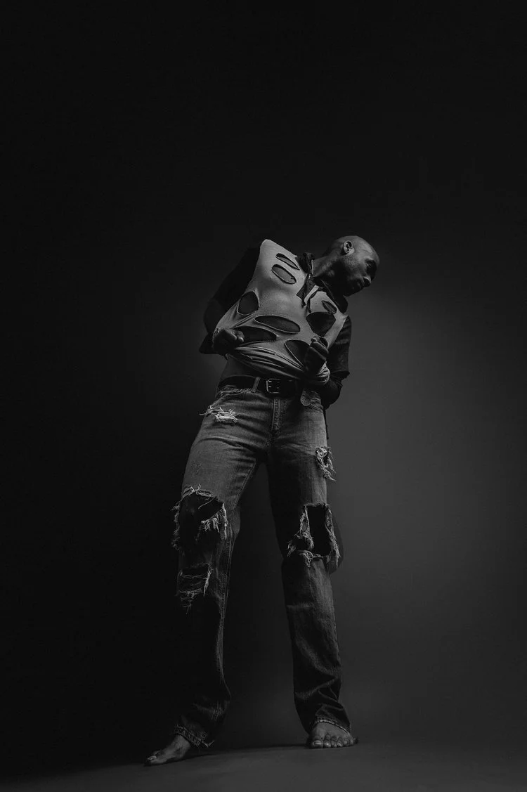 A person standing in a dark studio, wearing a torn hoodie and ripped jeans, with their head tilted sideways and arms crossed.