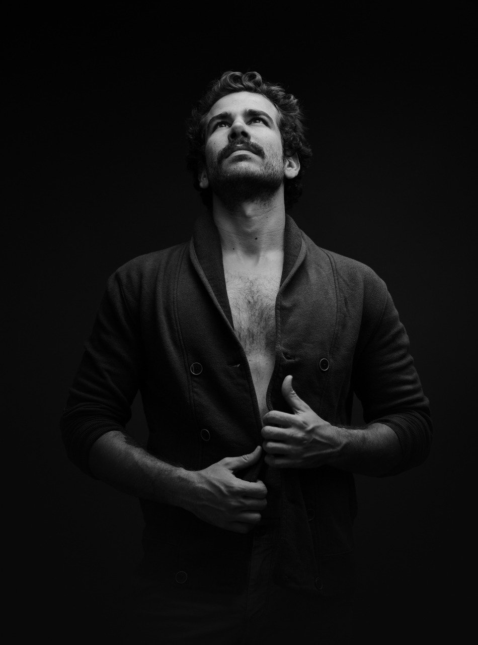 A black and white photo of a man with curly hair and a beard looking up with a serious expression, holding a partially unbuttoned jacket.