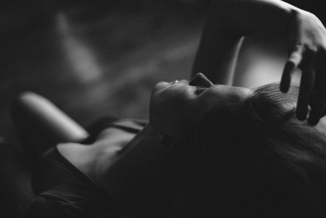 A woman lying down on her back, with her eyes closed, in a dimly lit setting, possibly resting or sleeping.