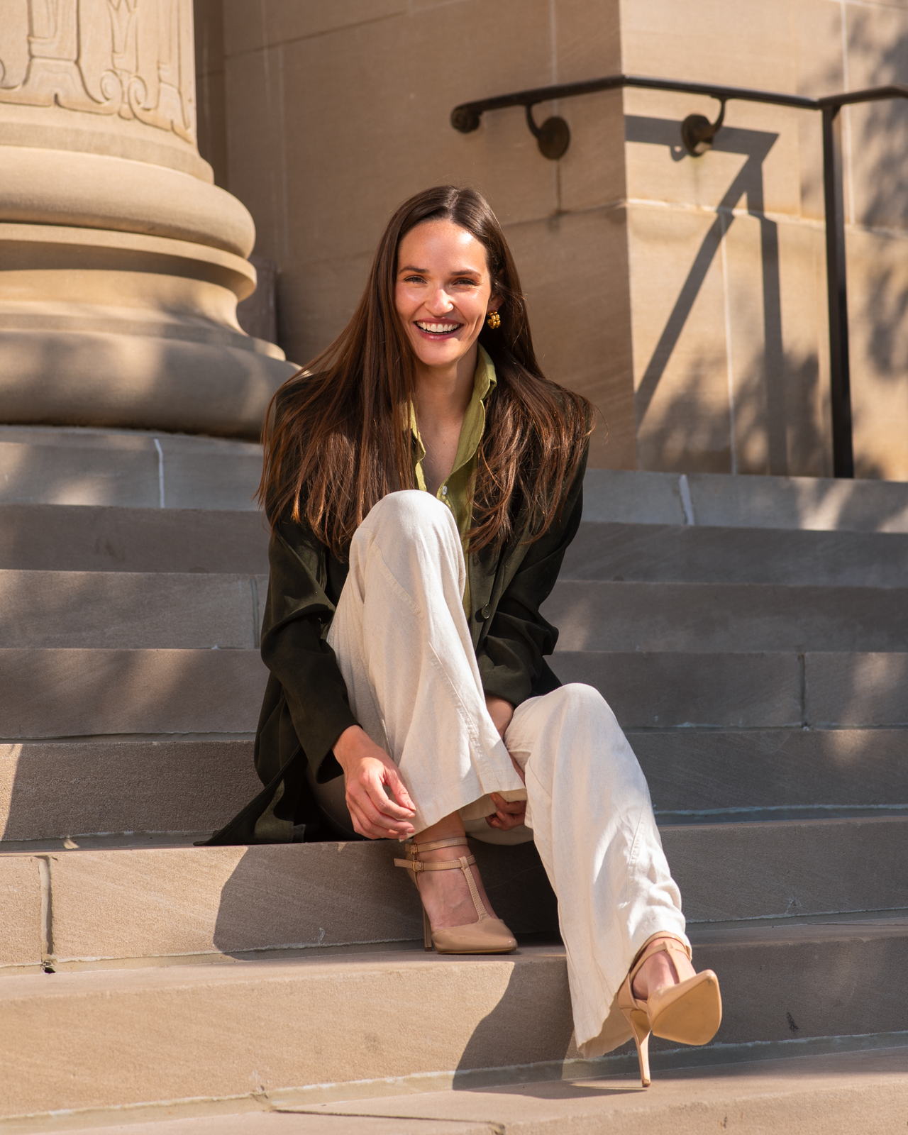 A woman with long brown hair sitting on steps outside a building, smiling, dressed in cream-colored pants, beige high heels, an olive green shirt, and a black jacket.