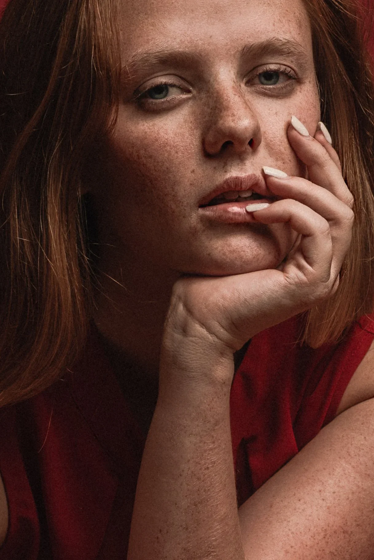 Close-up of a woman with red hair, freckles, and blue eyes, resting her chin on her hand, with her fingers near her lips, wearing a red sleeveless top.