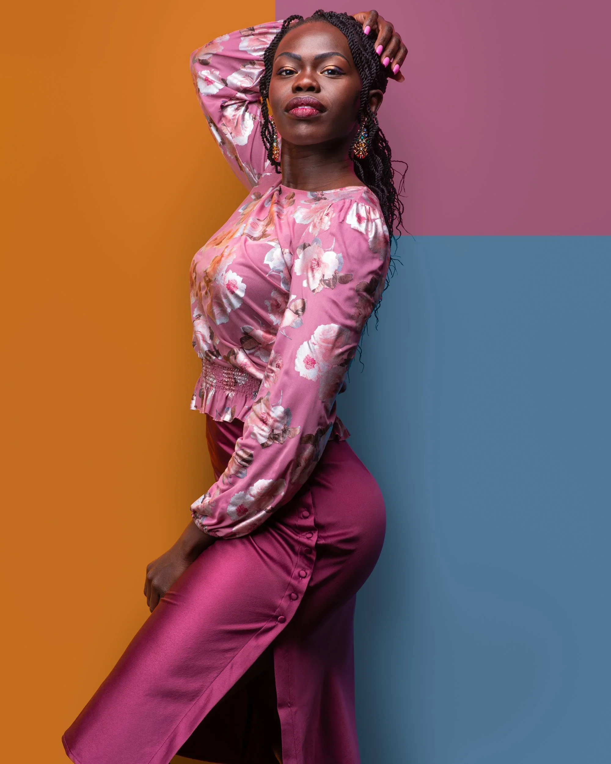 A woman with dark skin and braided hair posing against a colorful background divided into orange, purple, and blue sections. She is wearing a pink floral satin top and matching high-waisted pants, with colorful earrings and pink lipstick.