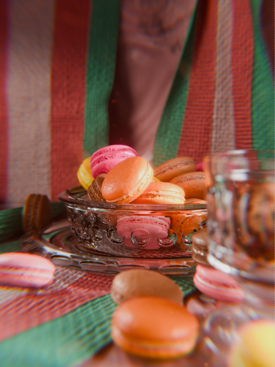 Colorful macarons in a glass dish with a blurred ceramic bowl and a colorful tablecloth in the background.
