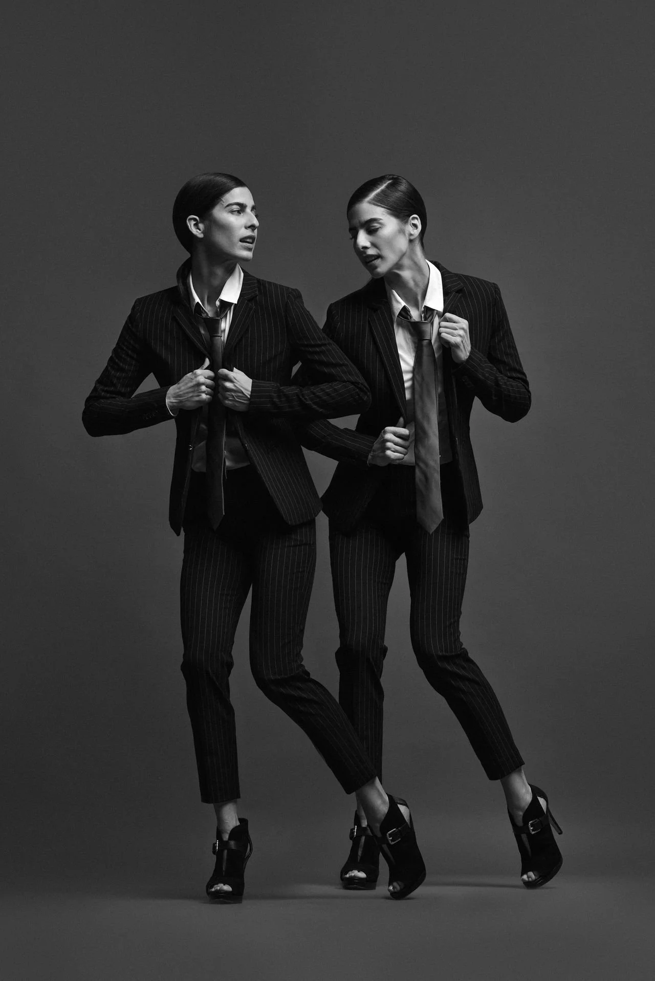 Two women in business suits with ties, standing close and engaging in conversation, one gesturing with her hand.
