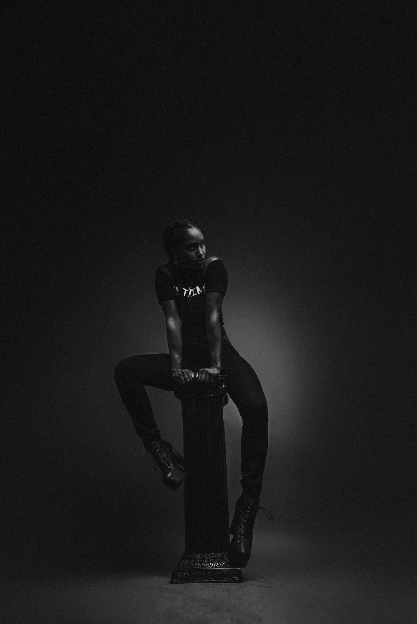 A woman in dark clothing and high-heeled boots sitting on a pedestal, in a black and white photograph with dramatic lighting.