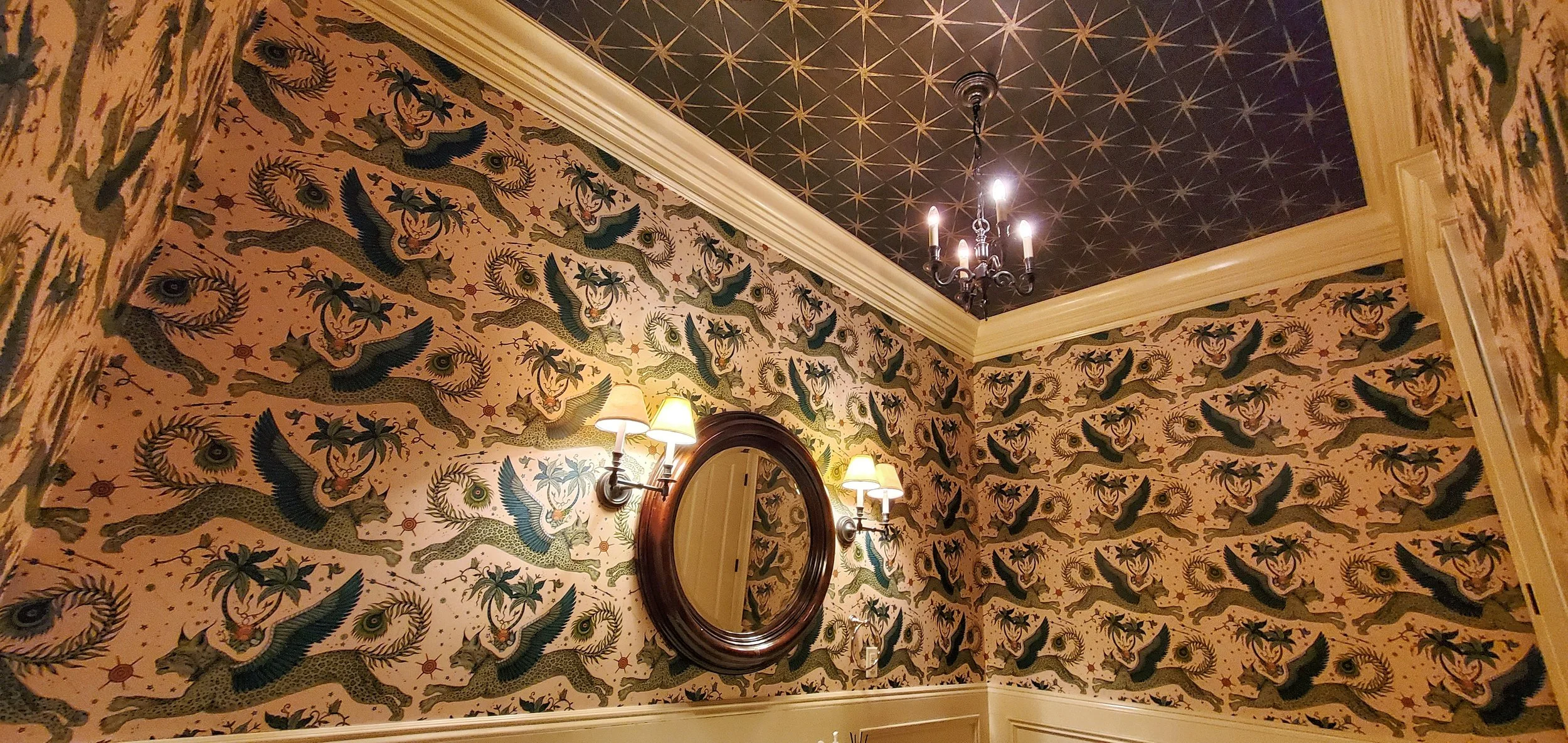 A Powder room with patterned wallpaper featuring Lynx flying griffins, palm trees, and staring eyes, with a evening star dark blue ceiling decorated with gold stars, a chandelier, and a wall-mounted mirror with a wooden frame.