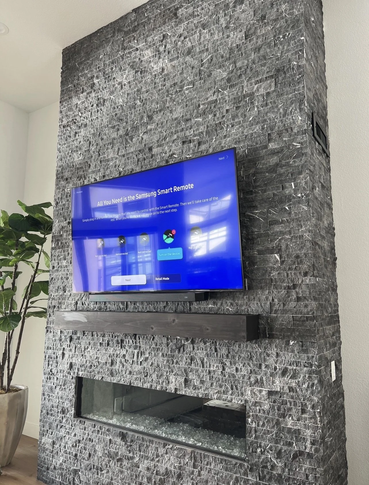 Living room TV mounted on a gray stone wall, displaying a setup screen for a Samsung smart remote. To the left is a tall potted plant with green leaves, and below the TV is a wooden shelf and a fireplace with glass stones inside.