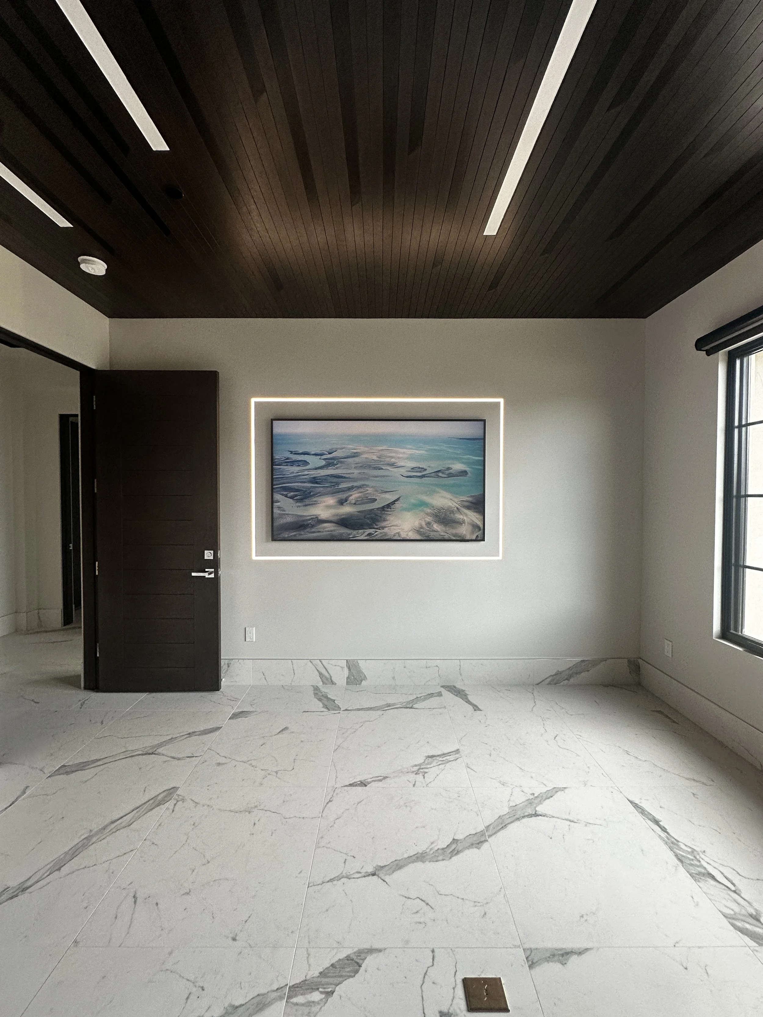 Empty modern room with marble floor, dark wooden ceiling with white accents, a large window on the right, a framed painting of an aerial landscape on the wall, and an open dark wooden door.