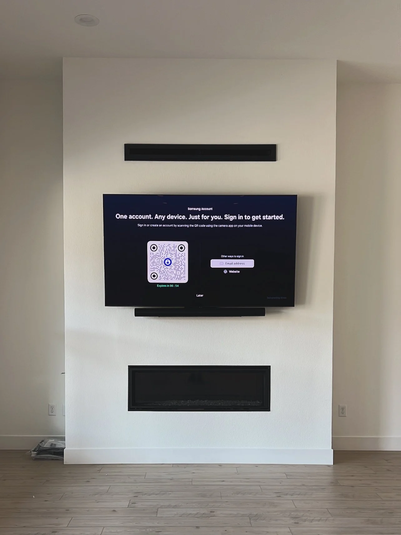 Living room with a wall-mounted flat-screen TV displaying a Samsung account sign-in screen, a television soundbar below the TV, and a modern fireplace below the soundbar in a room with light wooden flooring.