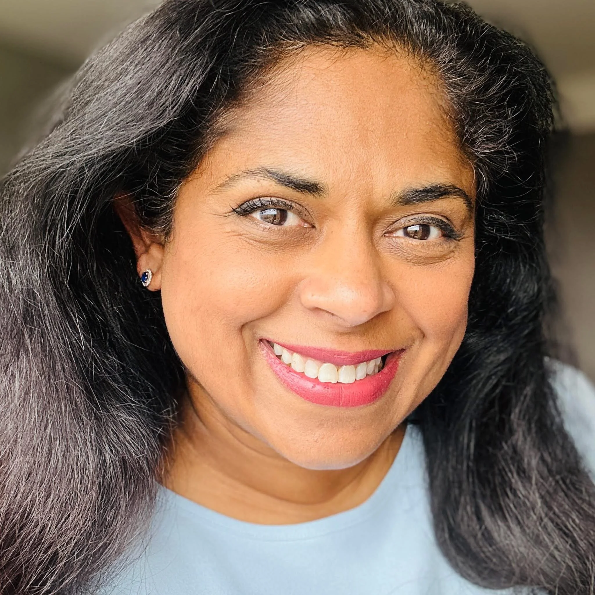 Close-up portrait of a smiling woman with dark hair, wearing earrings and a light-colored top.
