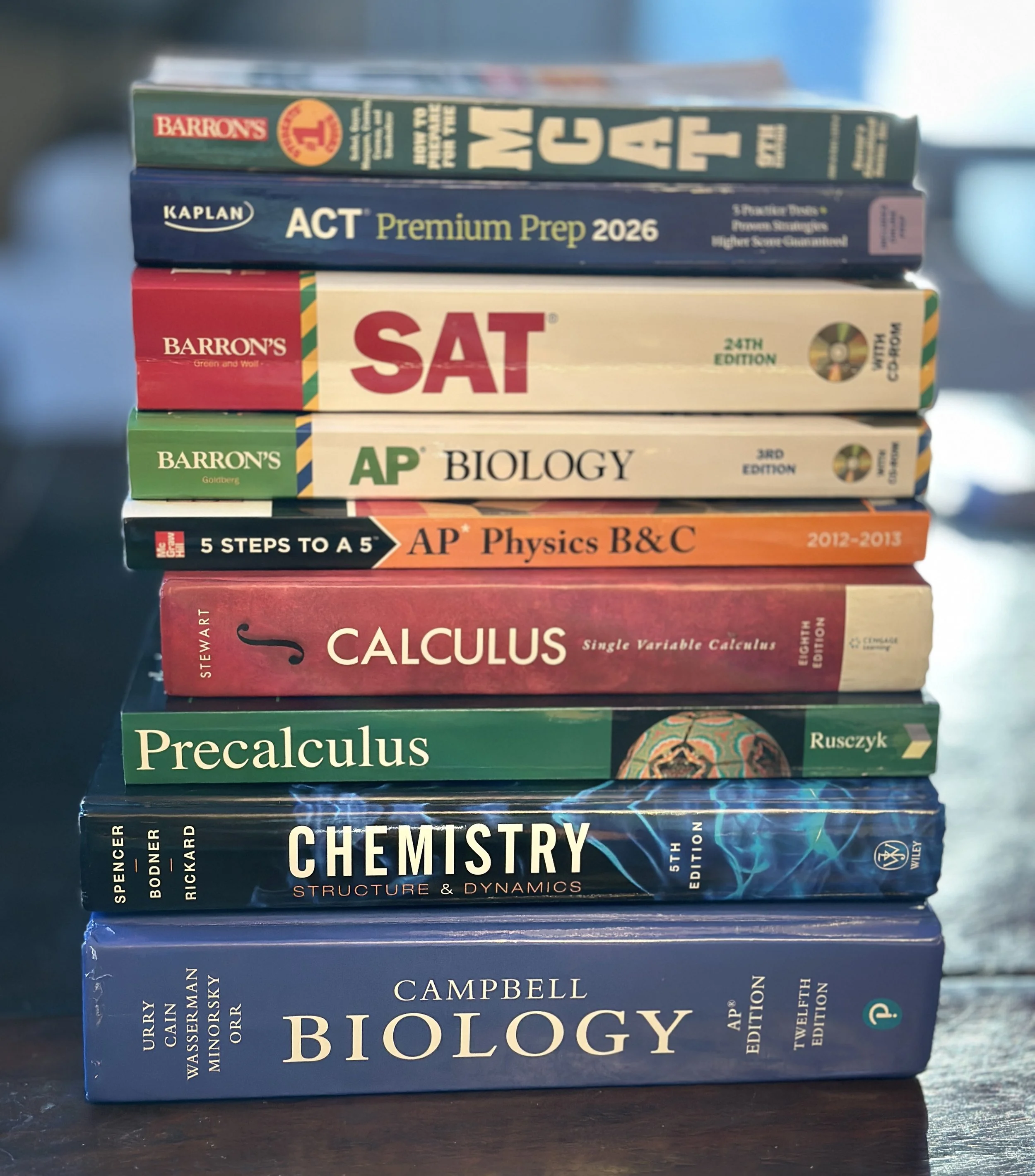 Stack of science and test preparation textbooks, including books on MGT, ACT, SAT, Biology, Physics, Calculus, Precalculus, Chemistry, and Biology.
