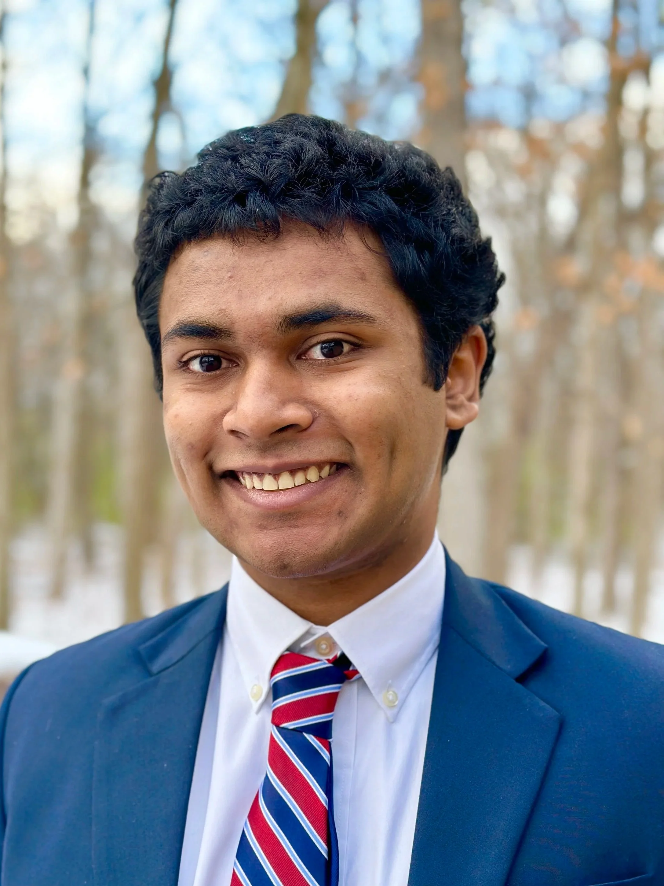 A young man with dark curly hair smiling outdoors, dressed in a navy blue suit, white shirt, and a red, white, and blue striped tie, with a background of leafless trees.