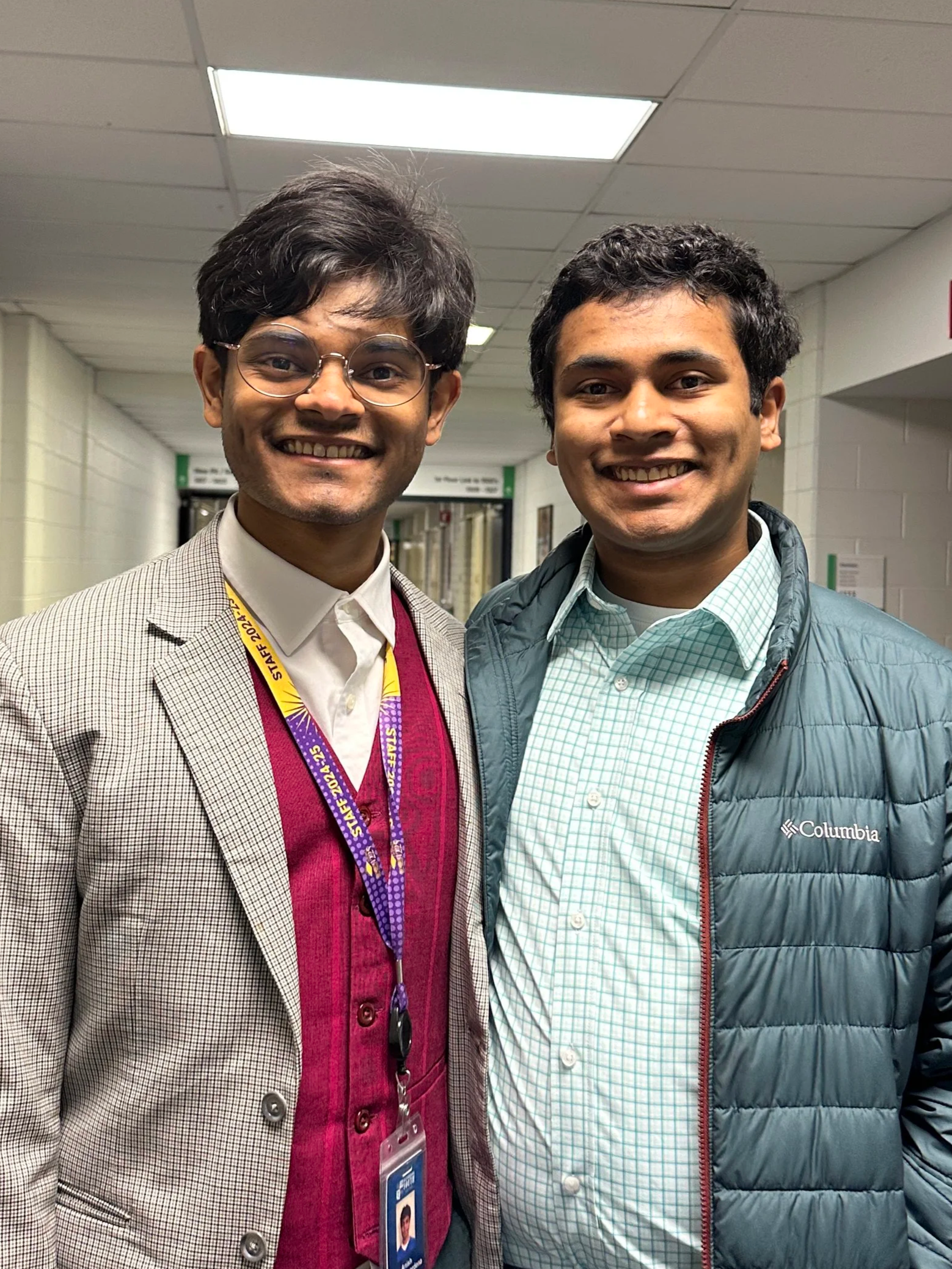 Two smiling men standing in a hallway, one wearing glasses and a checkered blazer, the other in a green plaid shirt and Columbia jacket.
