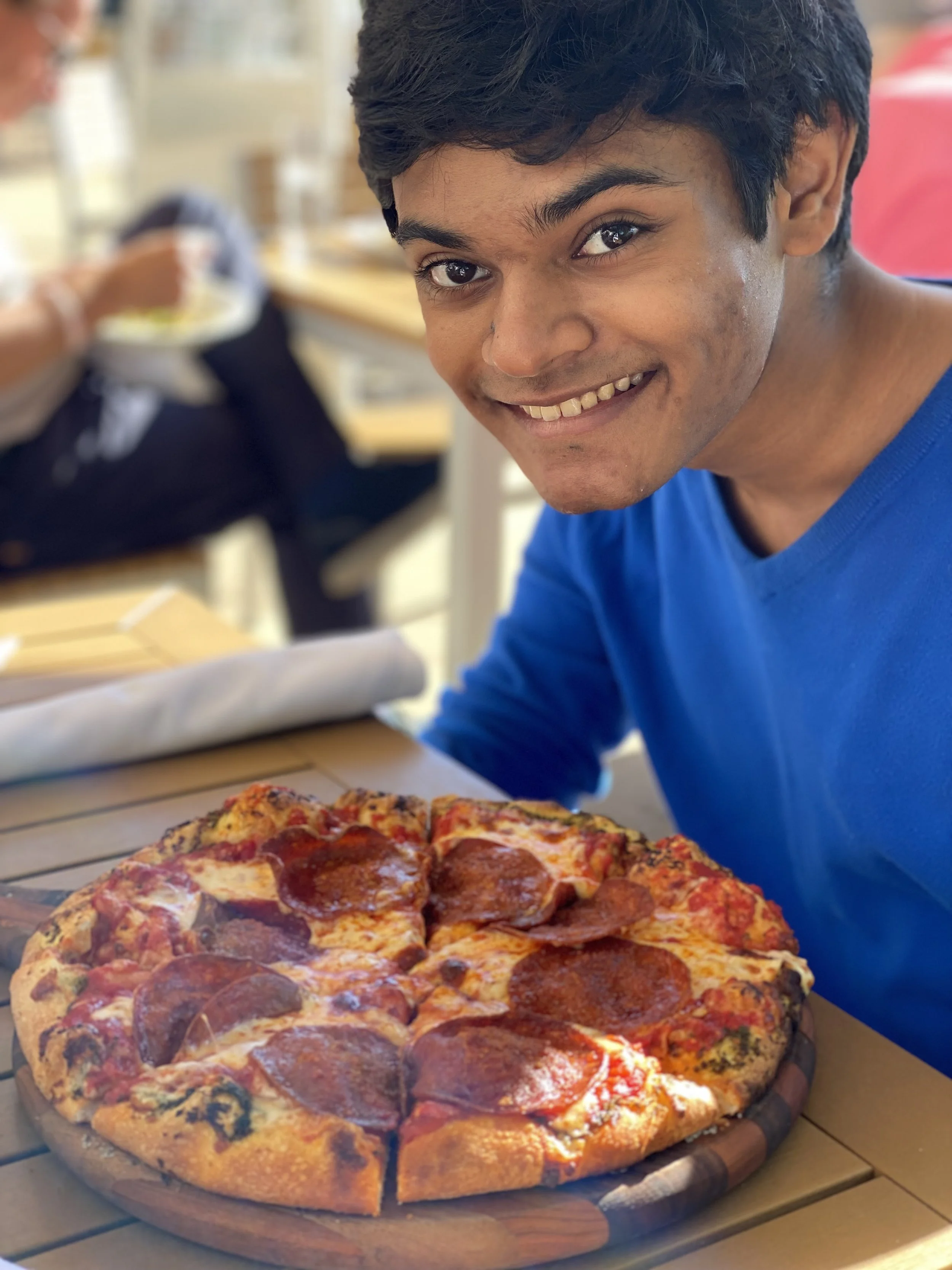 Young man smiling at a pizza with pepperoni slices on a wooden table.