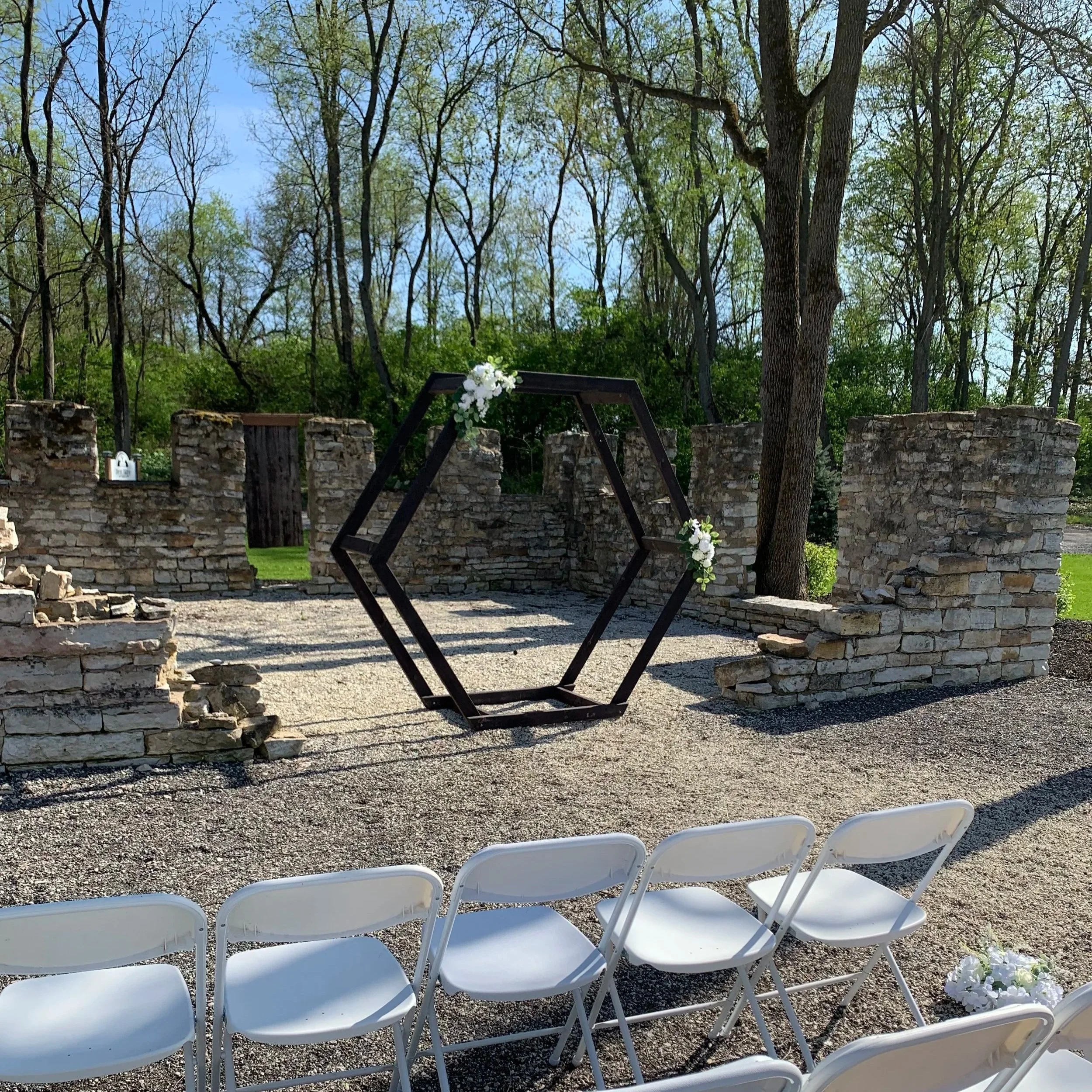 Outdoor wedding ceremony setup with white chairs, a geometric metal arch decorated with white flowers, and a stone wall backdrop surrounded by trees.