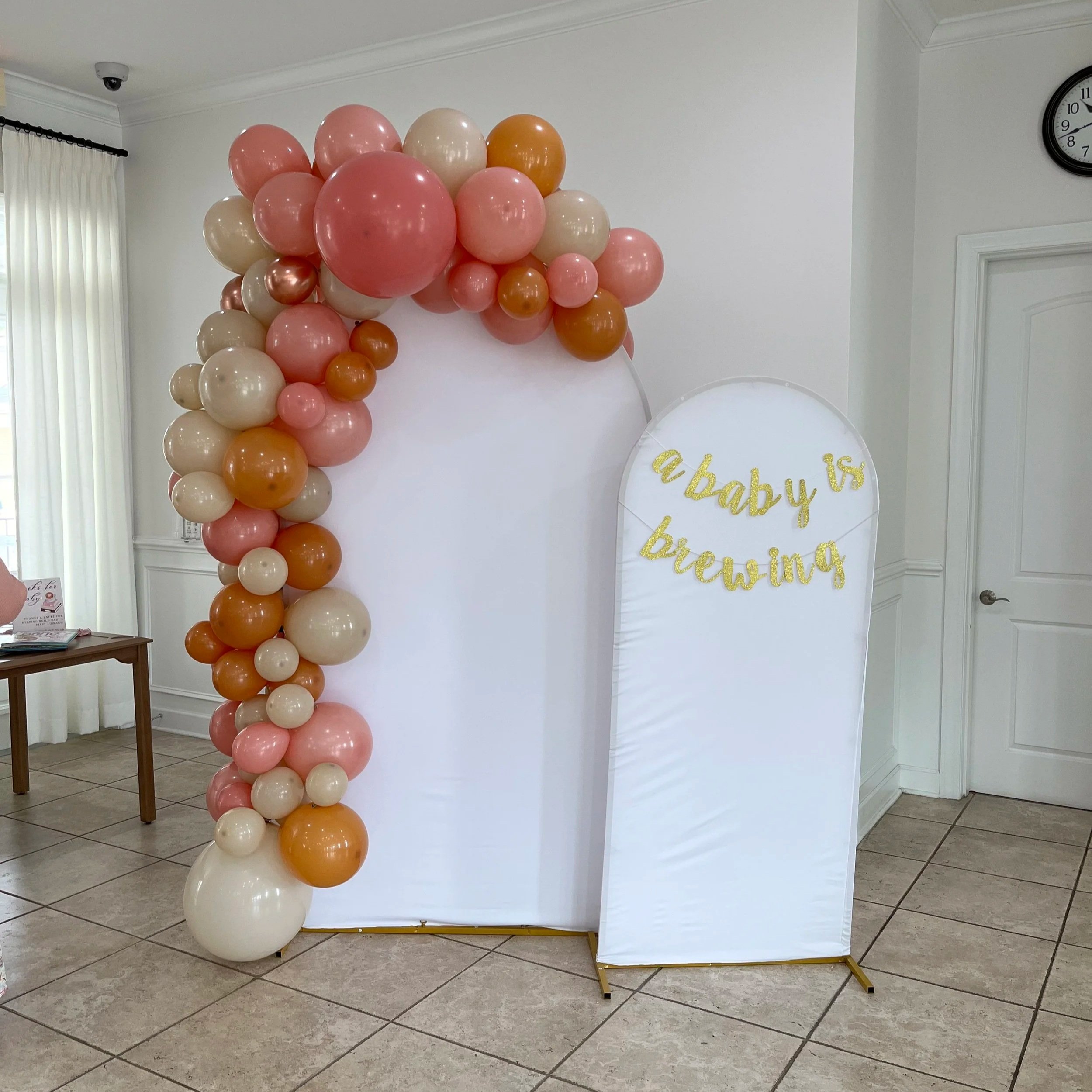 Decorative balloon arch with pink, cream, and orange balloons and a white banner with gold glitter text that reads 'a baby is brewing' in a room with tiled floor and white walls.