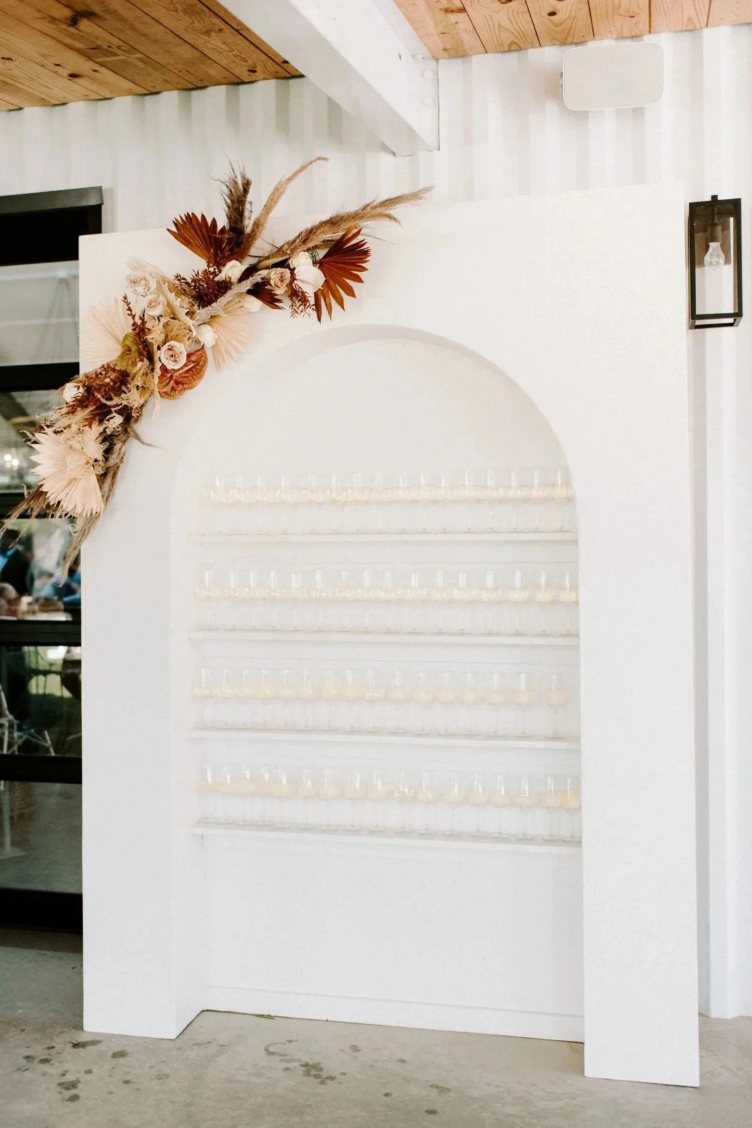 A white display wall with multiple rows of small clear glasses, decorated with a floral arrangement of dried flowers and leaves in shades of beige, brown, and rust, positioned in the top left corner.