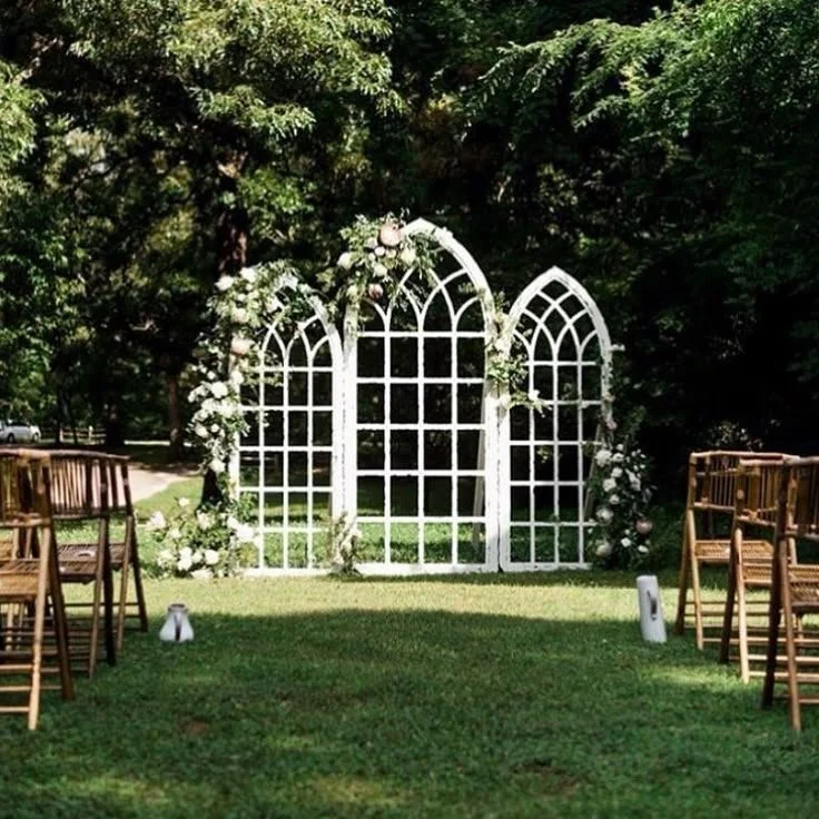Wedding ceremony setup outdoors with a white arched floral backdrop and wooden chairs arranged on a grassy lawn.