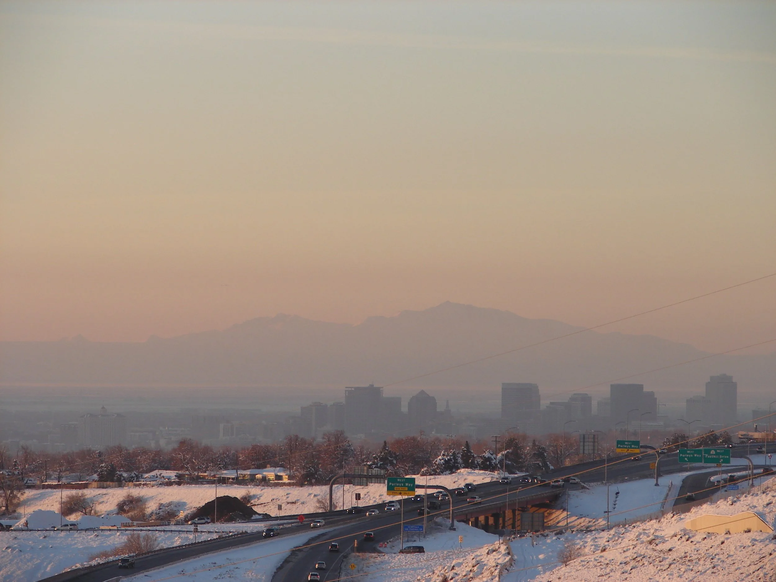 Snow-covered highway with cars and city skyline in the distance during sunset