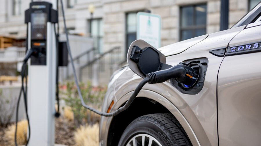 Close-up of an electric car plugged into a charging station, with a building and plants in the background.