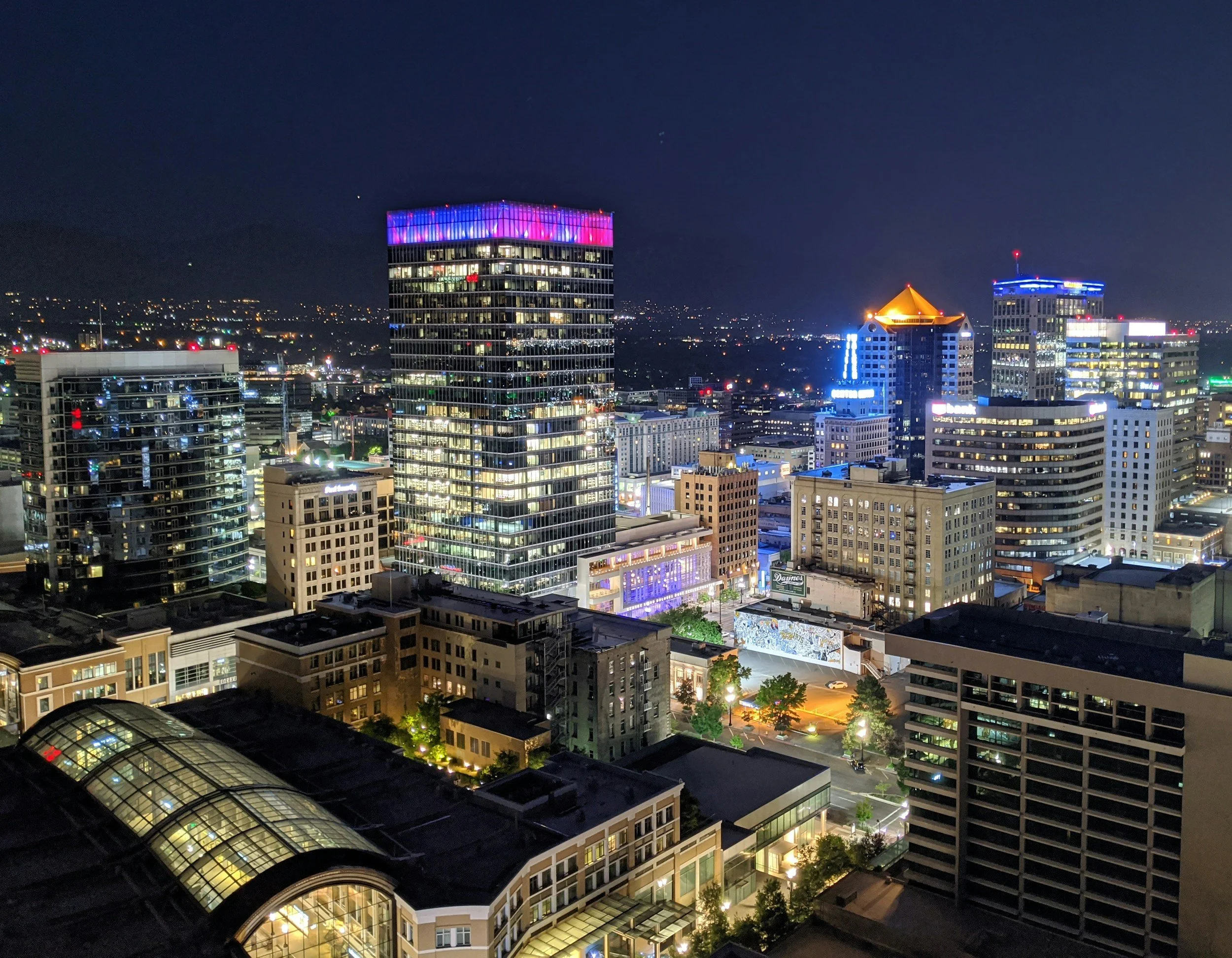 Nighttime cityscape with illuminated skyscrapers, including one with purple and pink lights on its upper levels, and streets below with lights and minor traffic.