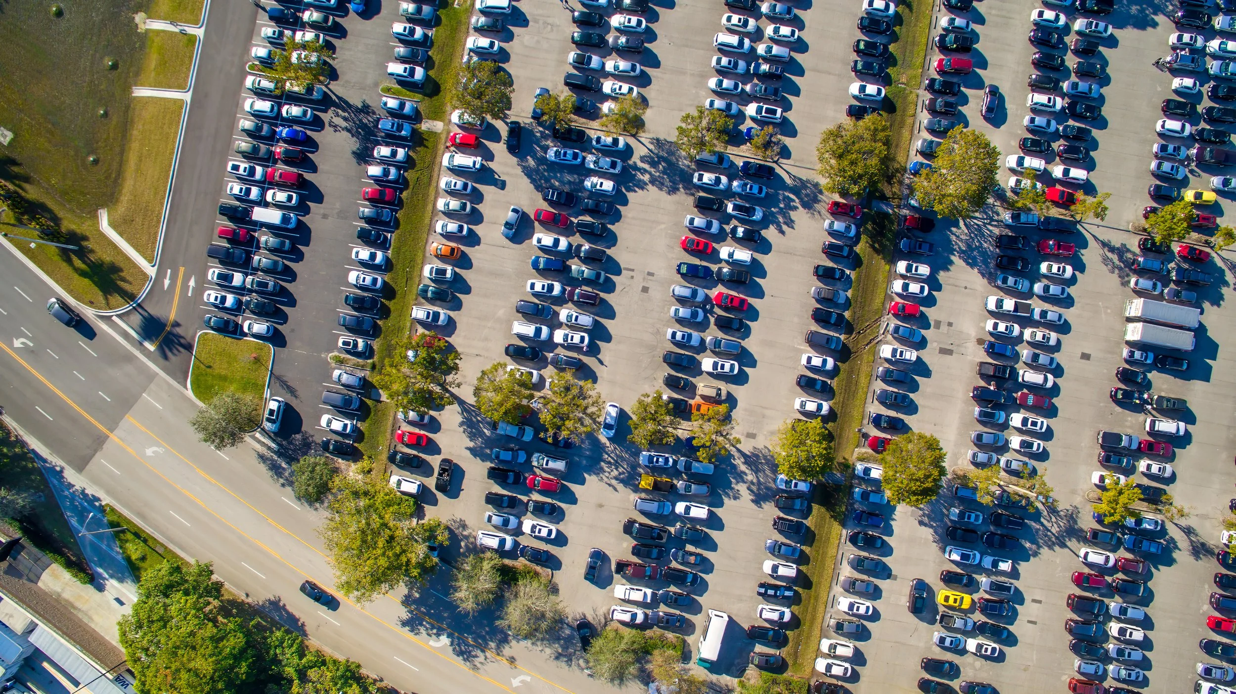 An aerial view of a large parking lot filled with numerous parked cars, some trees scattered throughout, and surrounding roads.