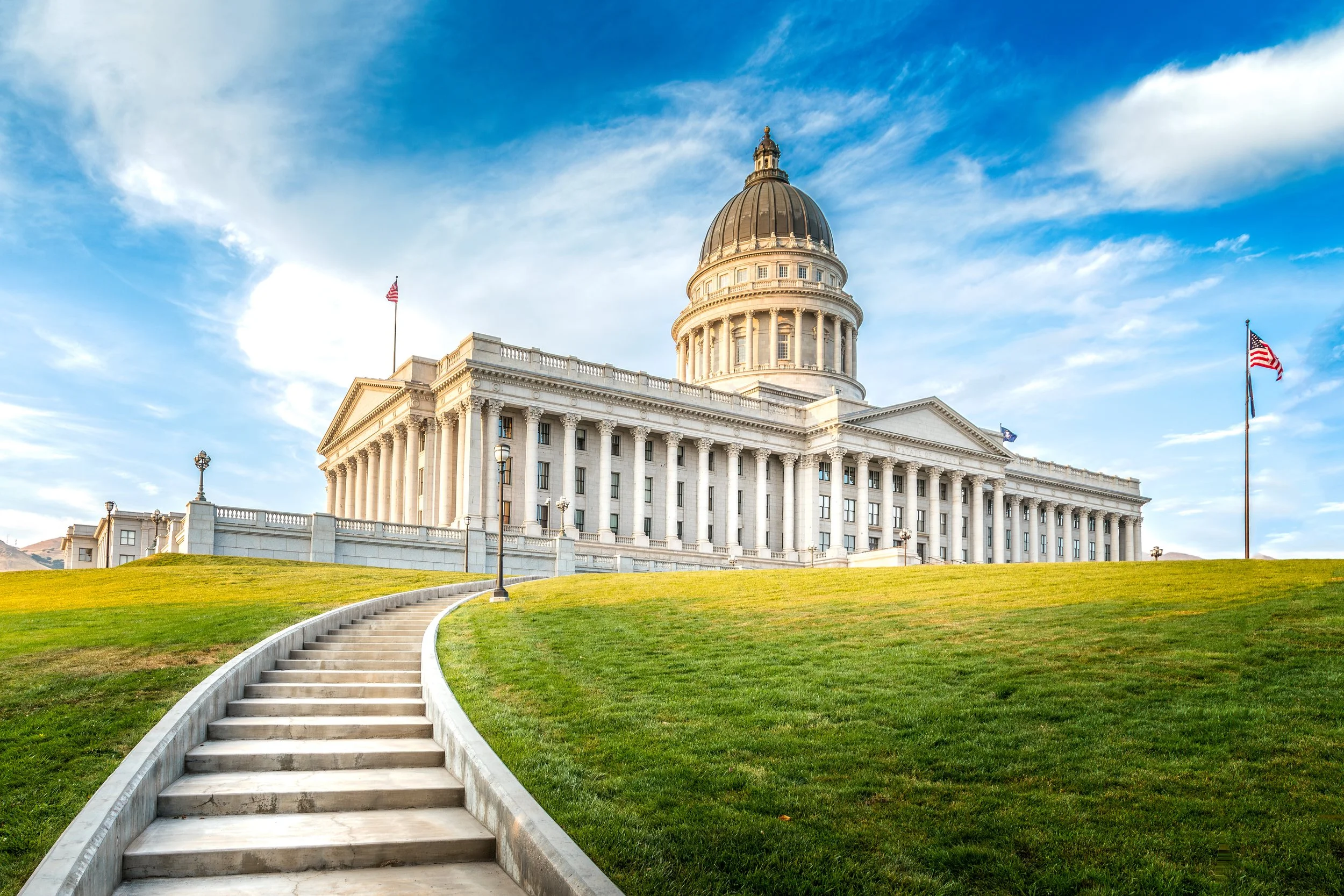 The image shows the United States Capitol building in Washington, D.C., with a green lawn and a curved pathway leading up to it, under a partly cloudy sky.