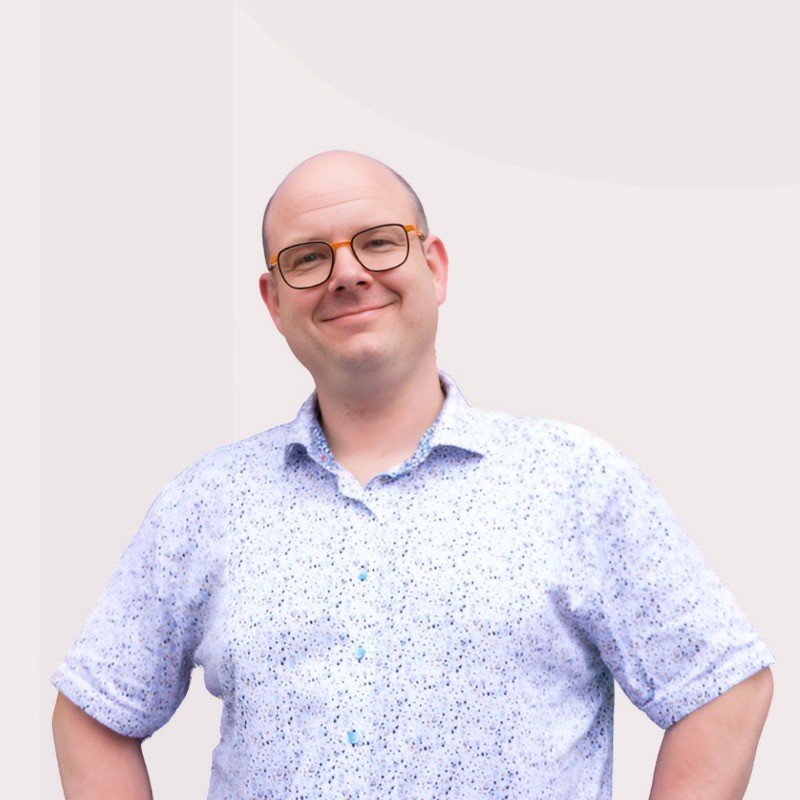 A smiling man with glasses, wearing a light-colored, patterned button-up shirt, standing against a light background.